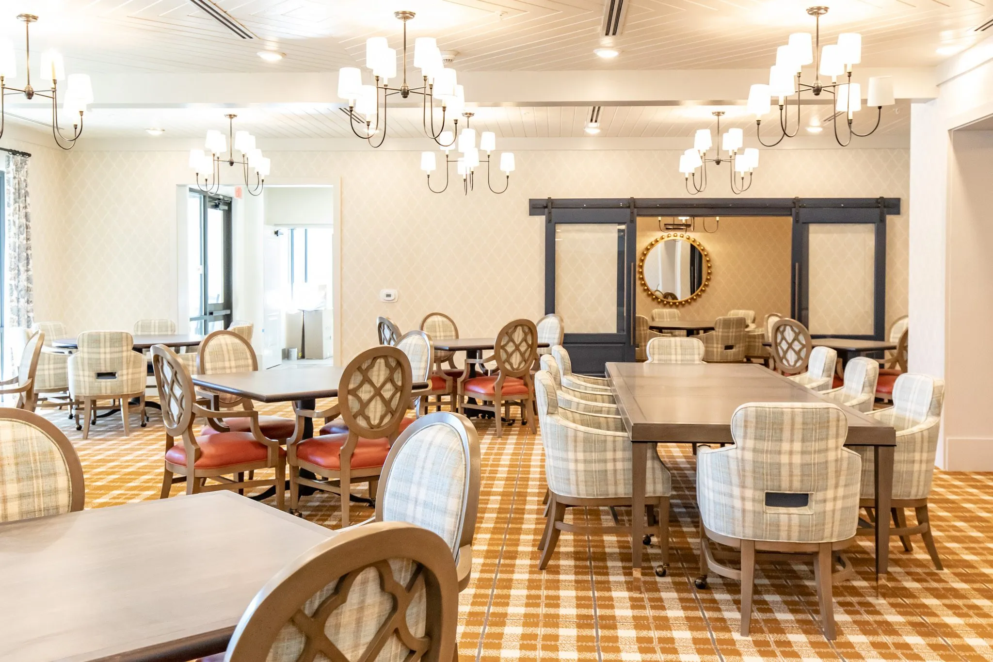 Senior living dining room toward the barn doors, a long table of plaid chairs and quatrefoil chairs under chandeliers