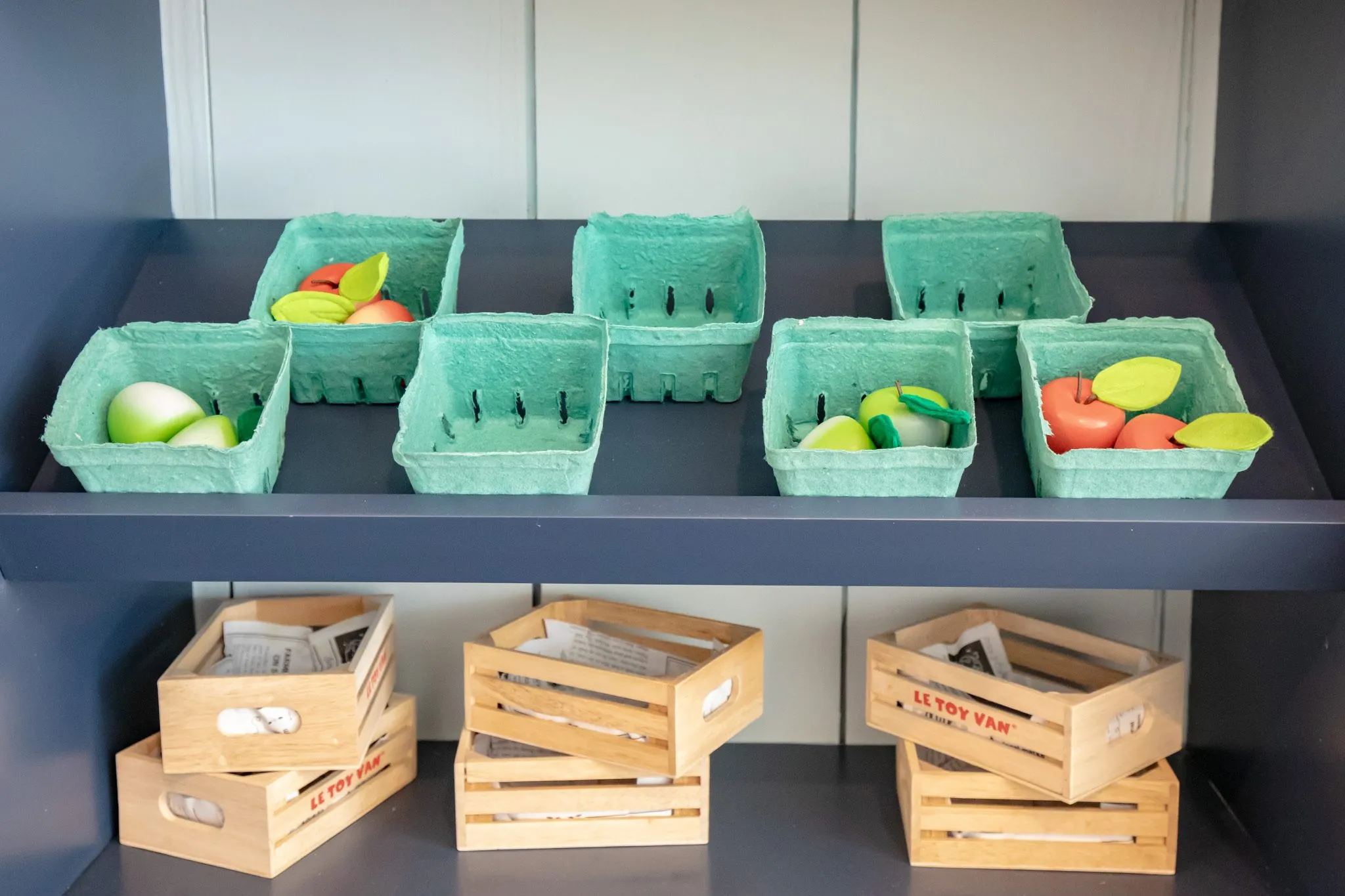 Senior living memory care market with green produce baskets of faux fruit on navy shelving above wooden crates