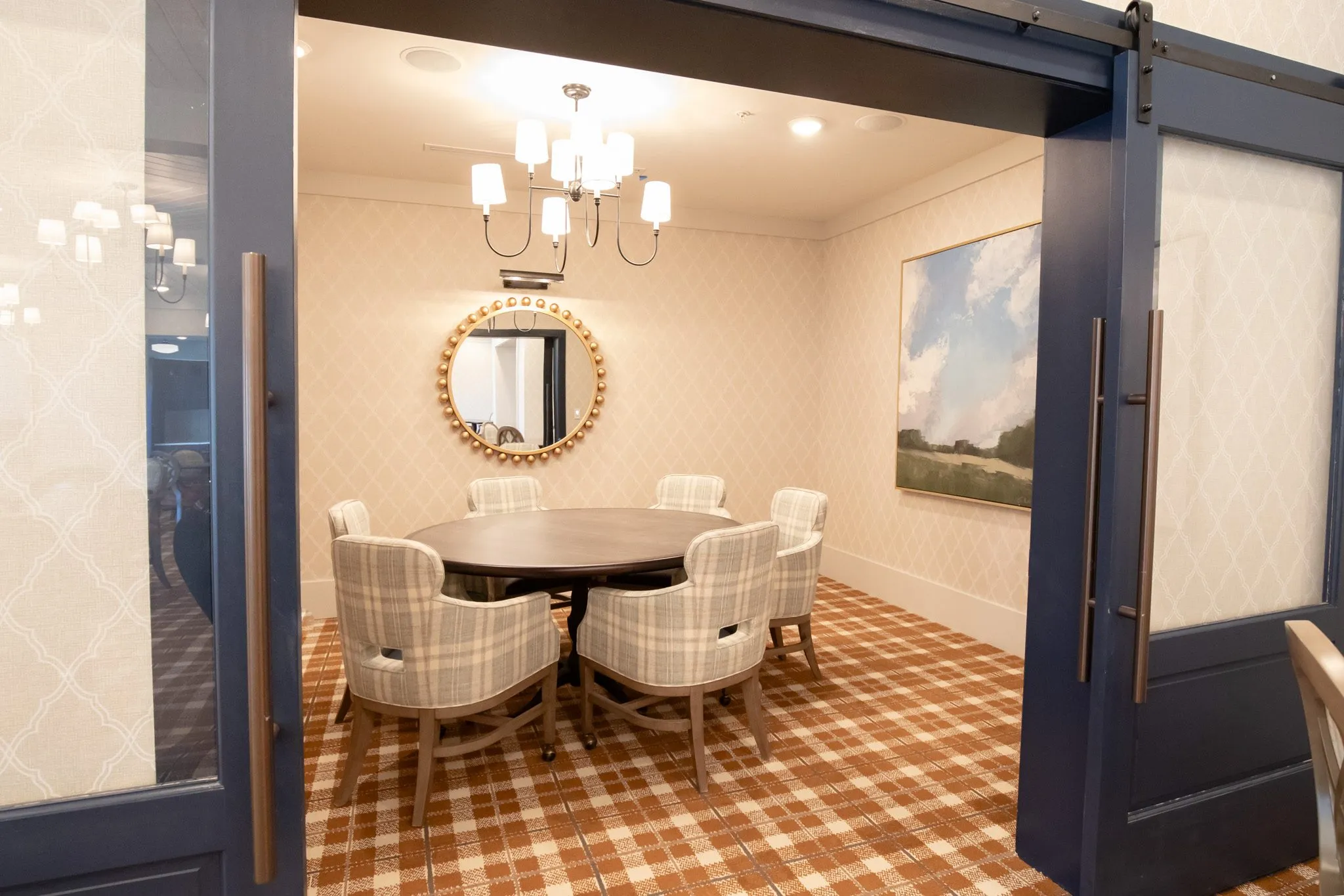Senior living private dining room with plaid chairs at a round table below a gilt beaded mirror, navy barn door alongside