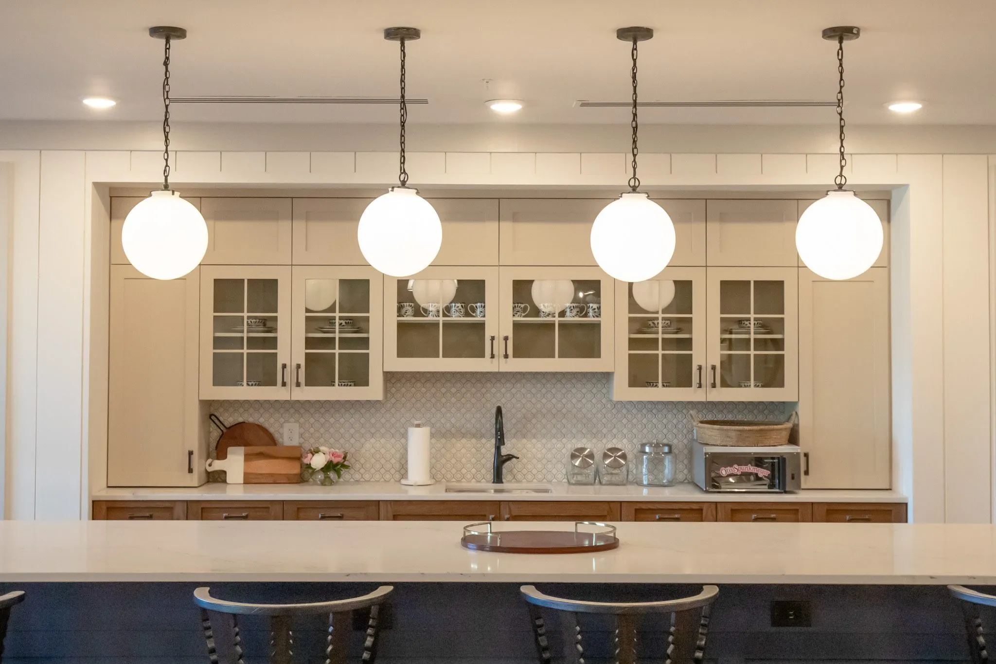 Senior living kitchen with four opal globe pendants over the island counter and glass-front cream cabinetry behind