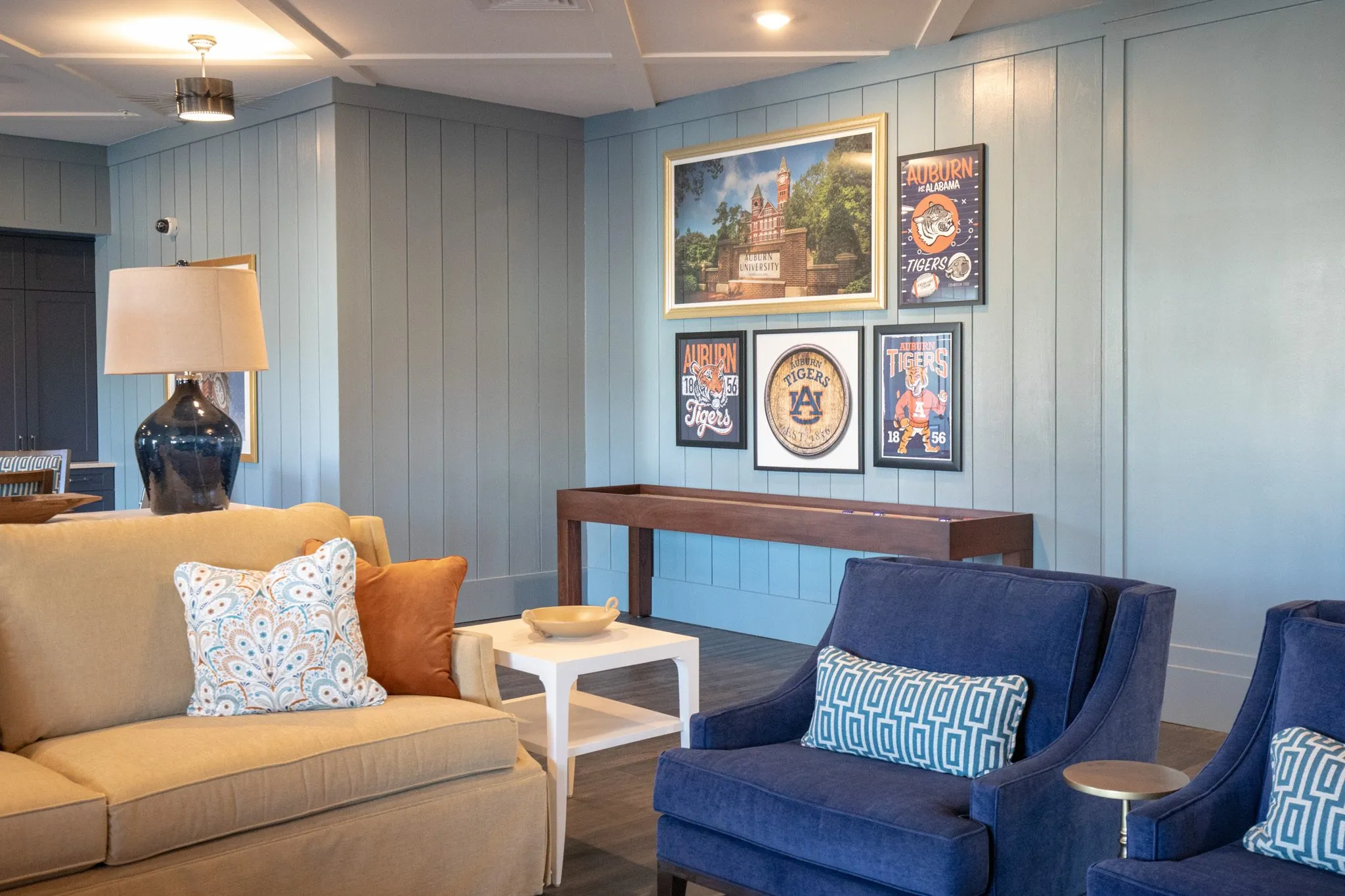 Senior living pub lounge with framed Auburn Tigers posters above a camel sofa and navy armchairs on pale blue shiplap