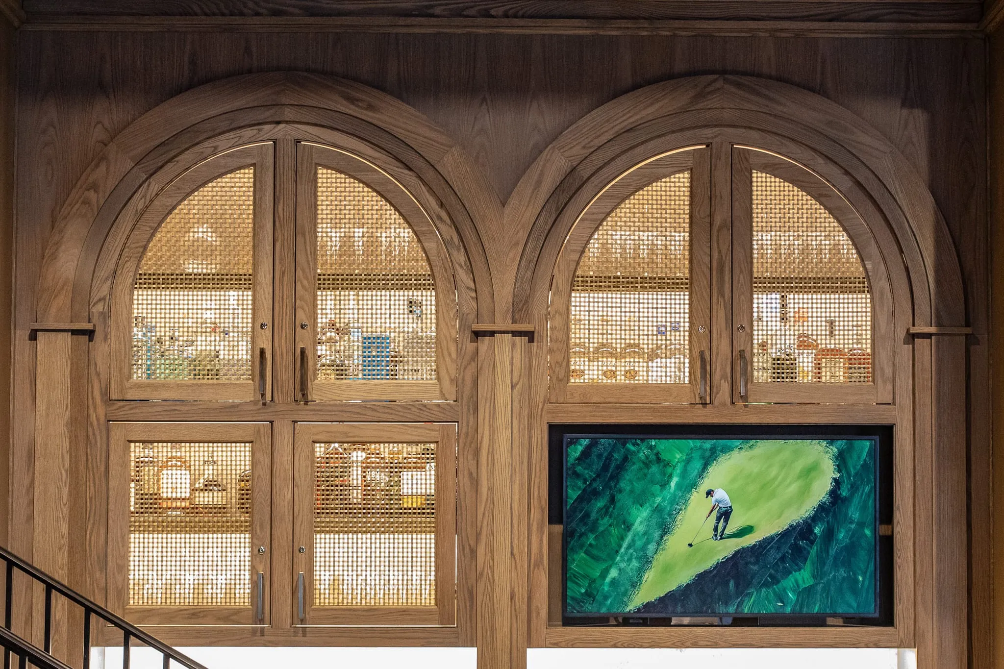 Private club interior detail: arched oak cabinetry with brass mesh doors framing a screen showing a golf course