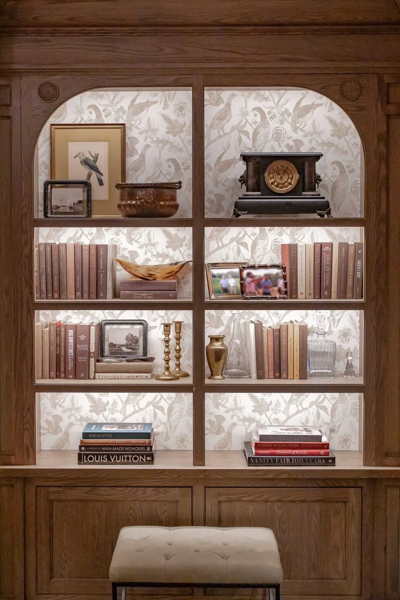 Private club interior detail: illuminated white oak bookcase with curated antiques and books on wallpapered shelves
