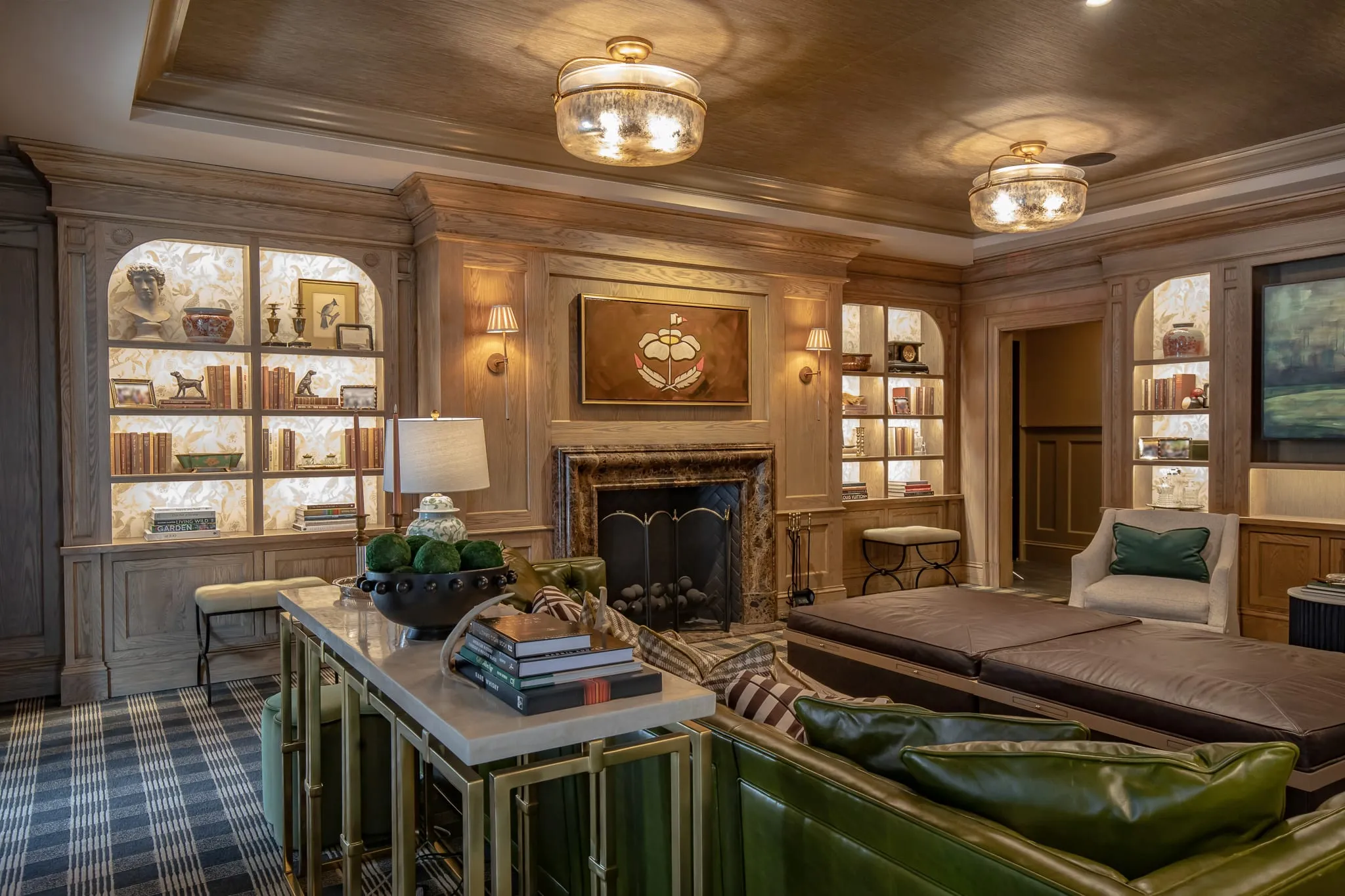 Private club library with a marble fireplace between lit open bookcases, a green leather sofa and tufted ottoman in front