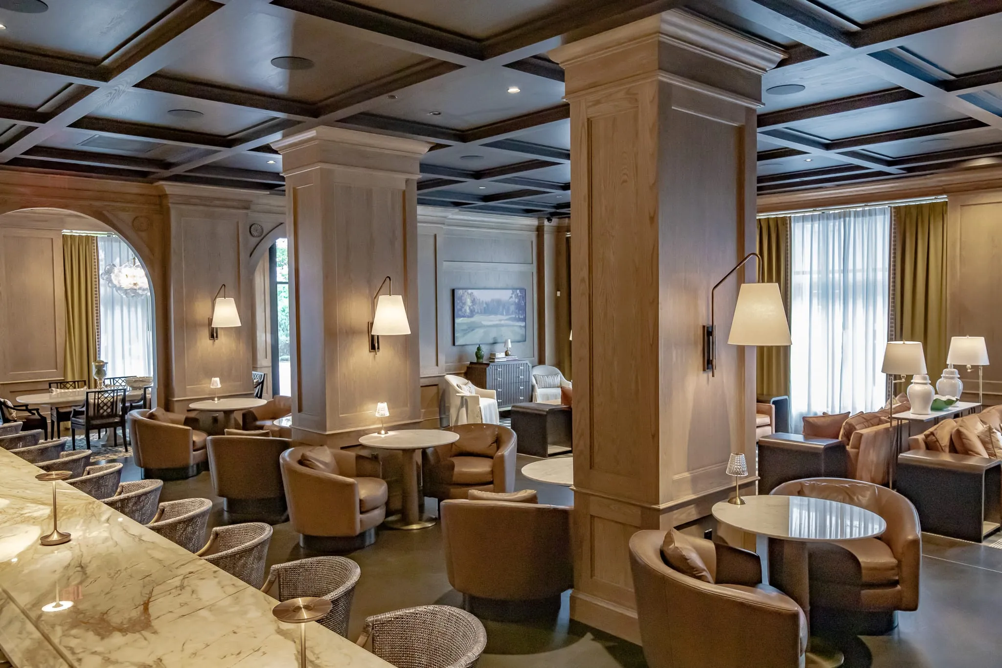 Club lounge with pale oak columns under a dark coffered ceiling, tan leather swivel chairs and marble cocktail tables