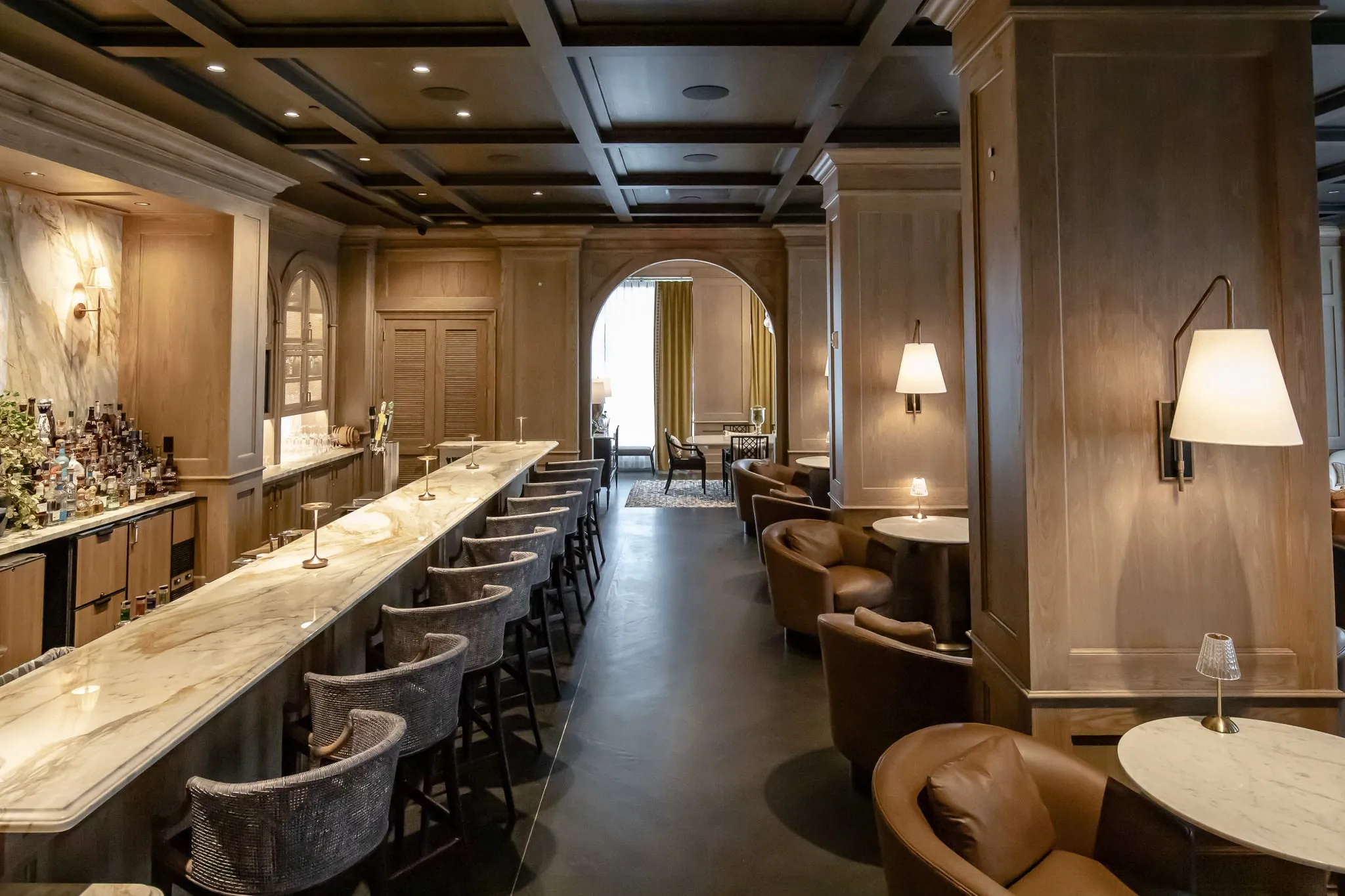 Private club bar with an uninterrupted marble slab counter and upholstered stools running its length to an arched doorway