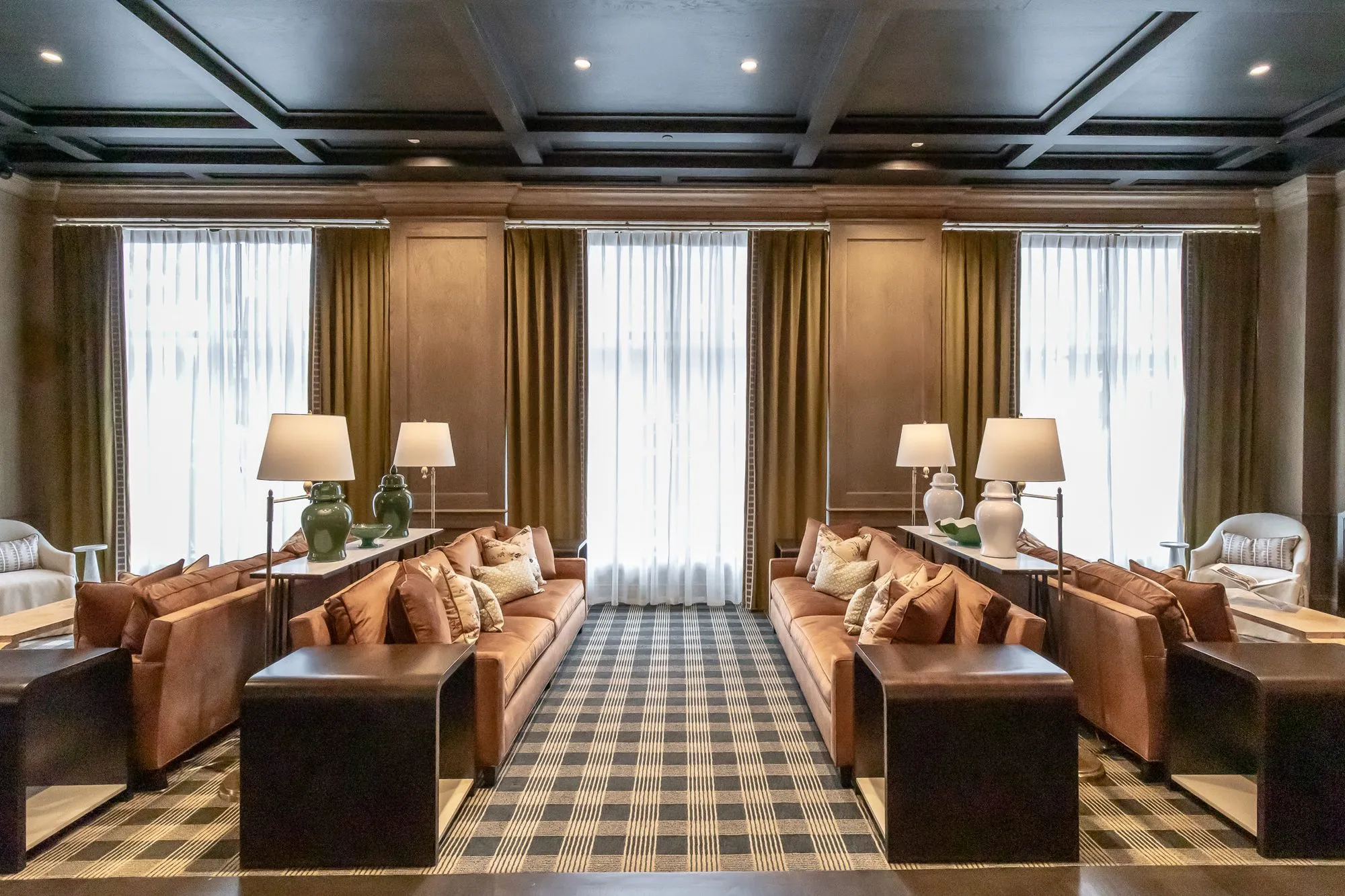 Private club lounge with cognac sofas facing across a checked carpet below tall curtained windows and a coffered ceiling