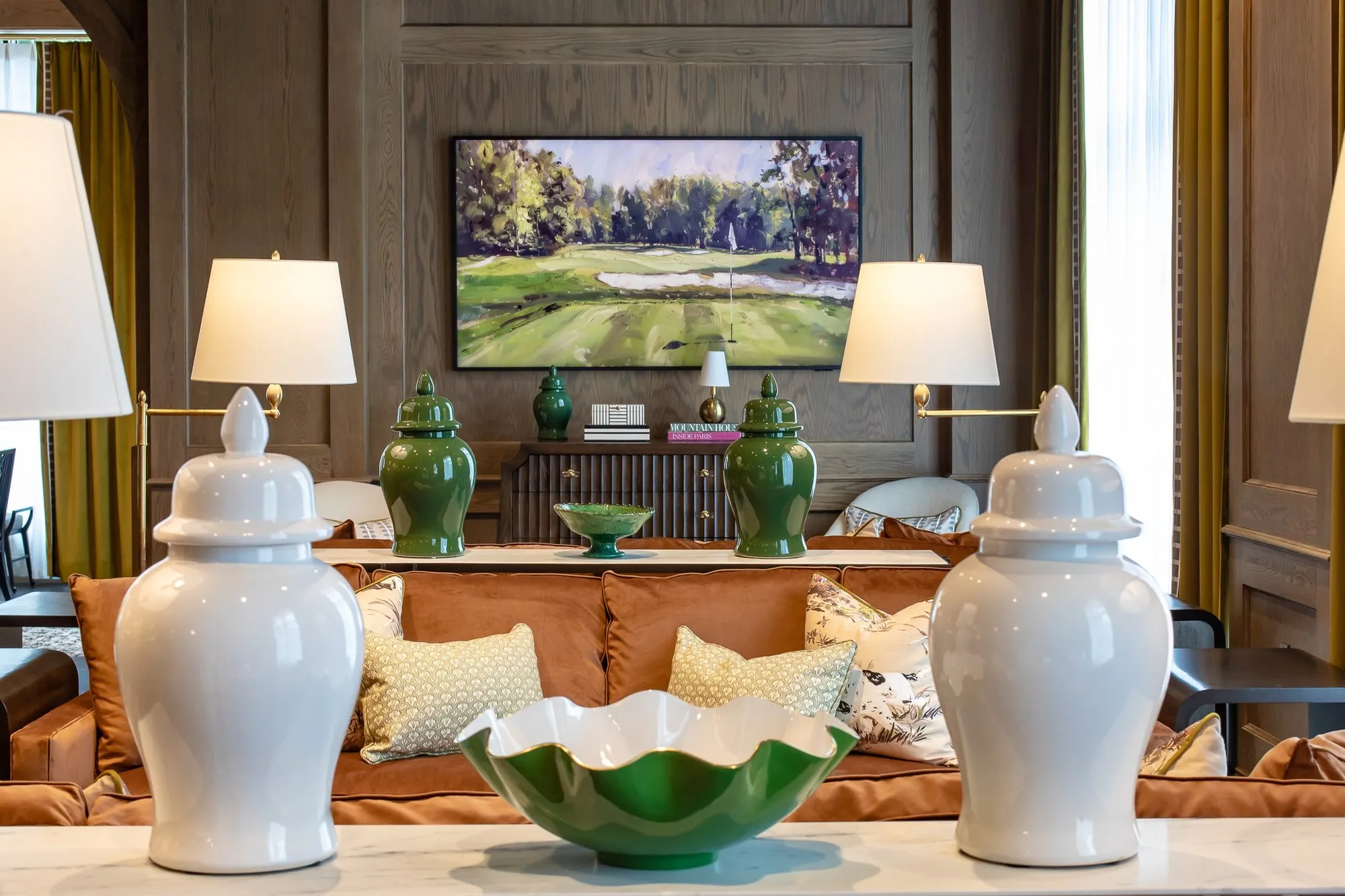 Private club interior detail: white ginger jars and a green scalloped bowl on a marble console, a golf course painting beyond