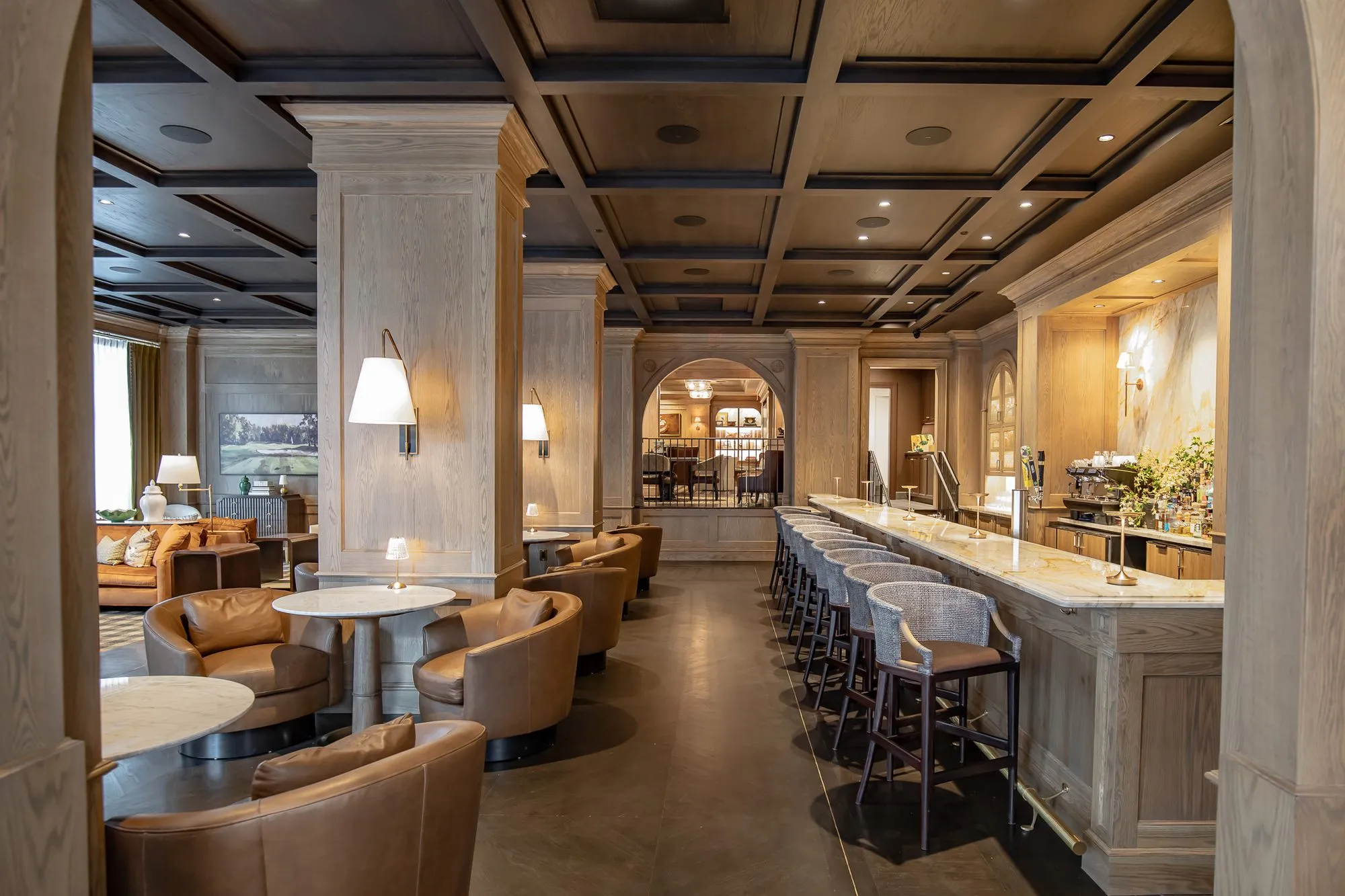 Private club bar lounge with leather tub chairs at marble tables beside the long marble bar under a coffered ceiling
