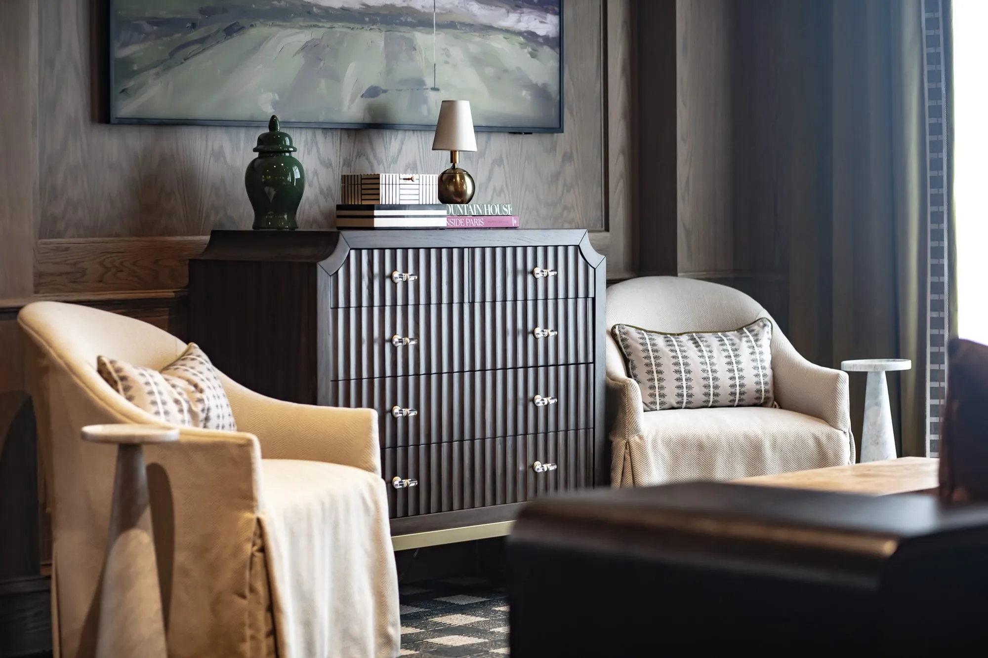 Private club interior detail: two slipcovered armchairs flanking a fluted grey chest below a landscape painting on oak