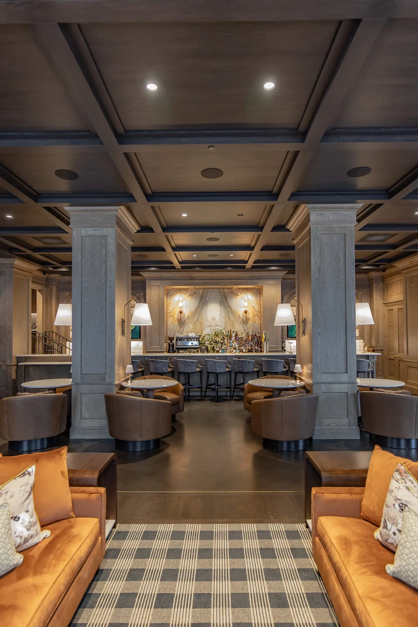 Private club bar lounge seen from between cognac sofas to the marble bar under the coffered ceiling