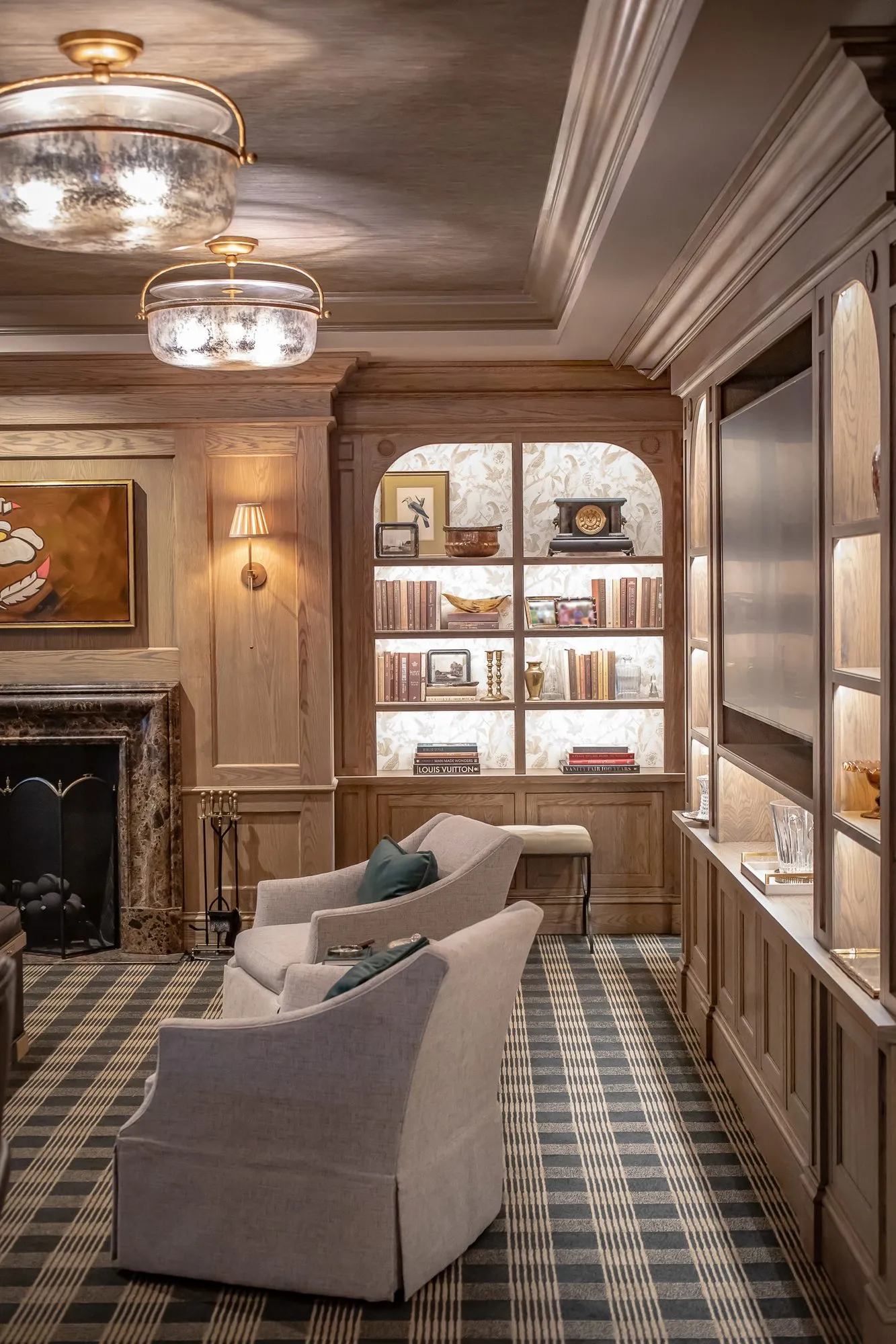 Private club library corner with skirted armchairs on a checked carpet beside a marble fireplace and lit bookcase