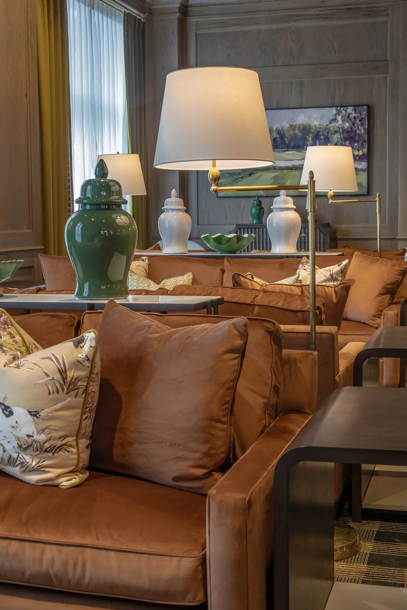 Private club interior detail: cognac velvet sofa with floral pillows, swing-arm floor lamps and a golf painting beyond