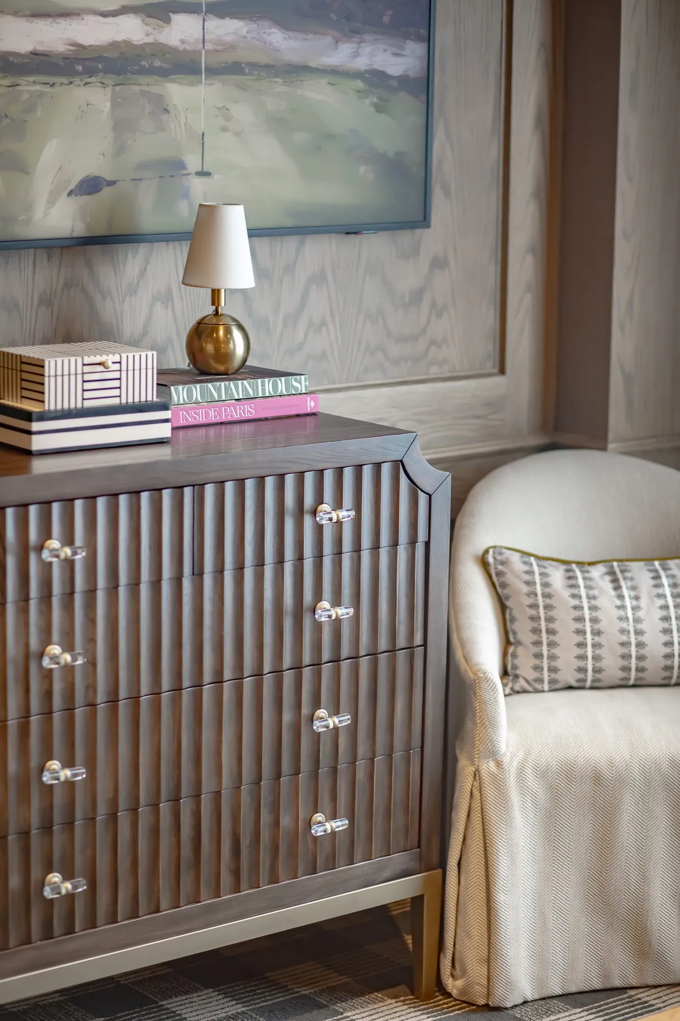 Private club interior detail: fluted grey chest with glass knobs, a brass lamp and books, a slipcovered chair beside it