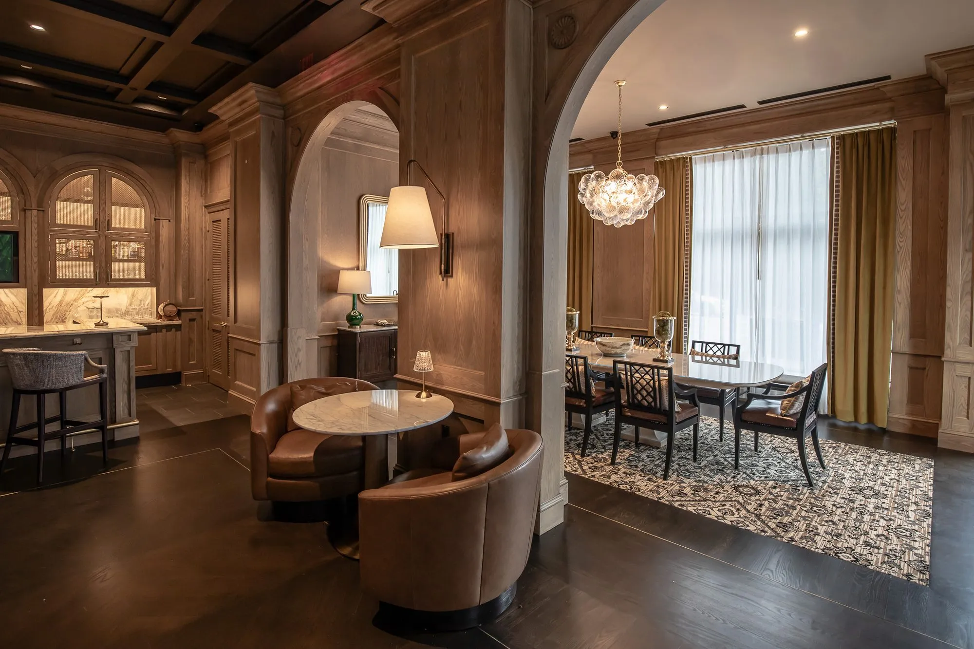 Private club lounge with leather tub chairs at a marble table between archways to the private dining room and marble bar