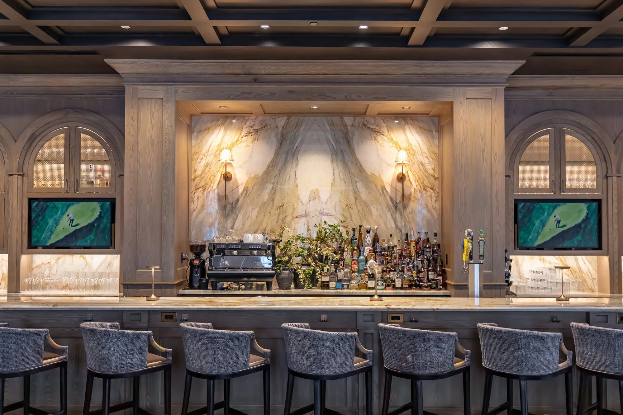 Private club bar back with a book-matched marble slab and bottles between arched glass cabinets, grey stools in front