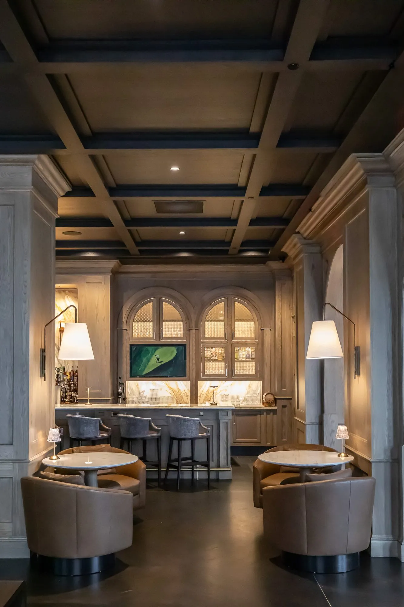 Private club bar seen between oak columns under the coffered ceiling, leather tub chairs at marble tables
