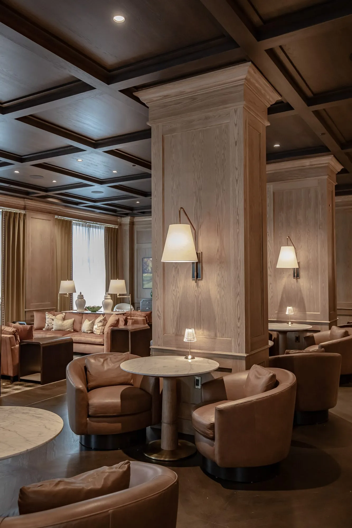 Private club lounge with leather tub chairs at marble tables by an oak column and linen sconce, sofas along the windows
