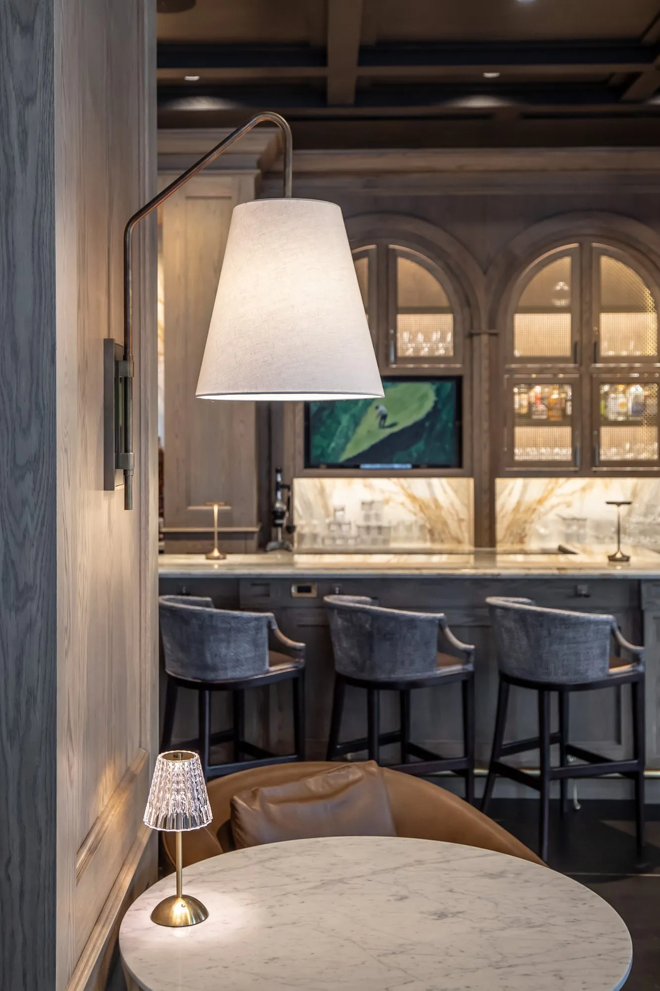 Private club bar with a swing-arm lamp over a marble cafe table, grey stools at the marble bar and arched cabinets beyond