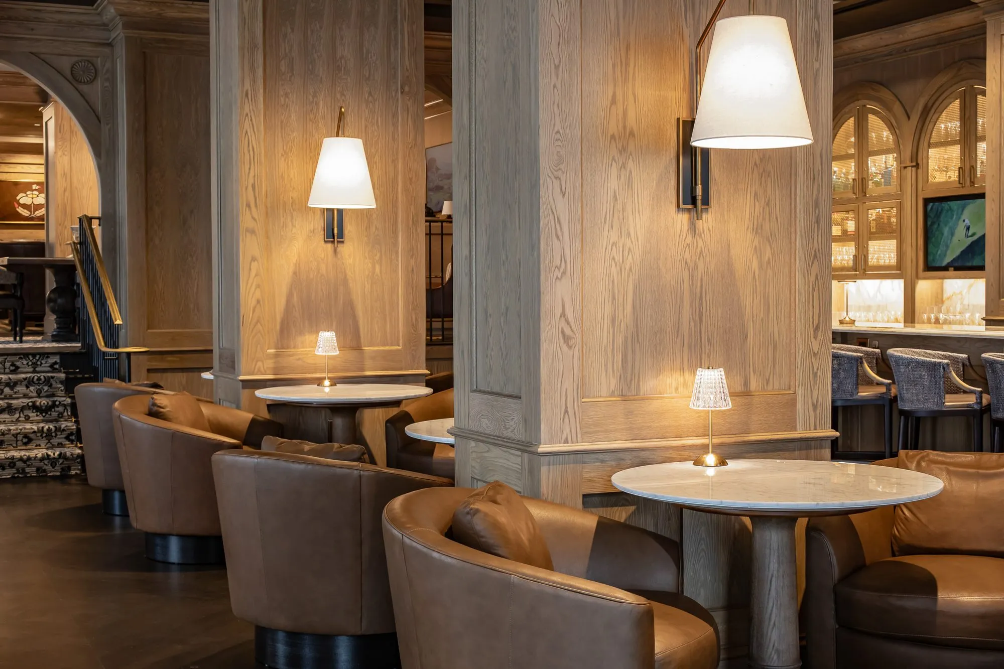Private club bar lounge with leather tub chairs at marble tables around oak columns with linen sconces, the patterned stair