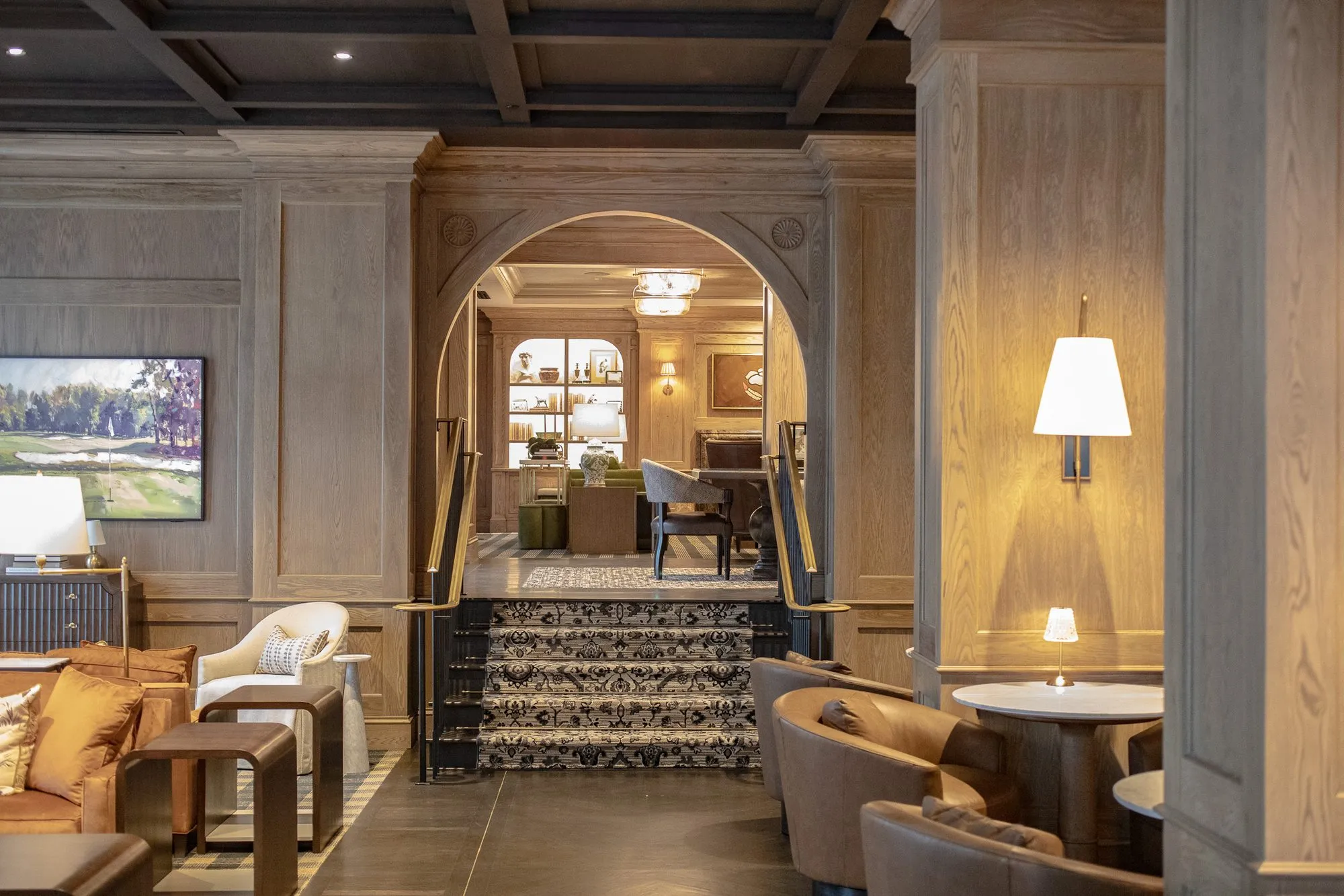 Private club lounge with leather tub chairs beside an oak arch and a patterned stair rising to a room with lit shelving