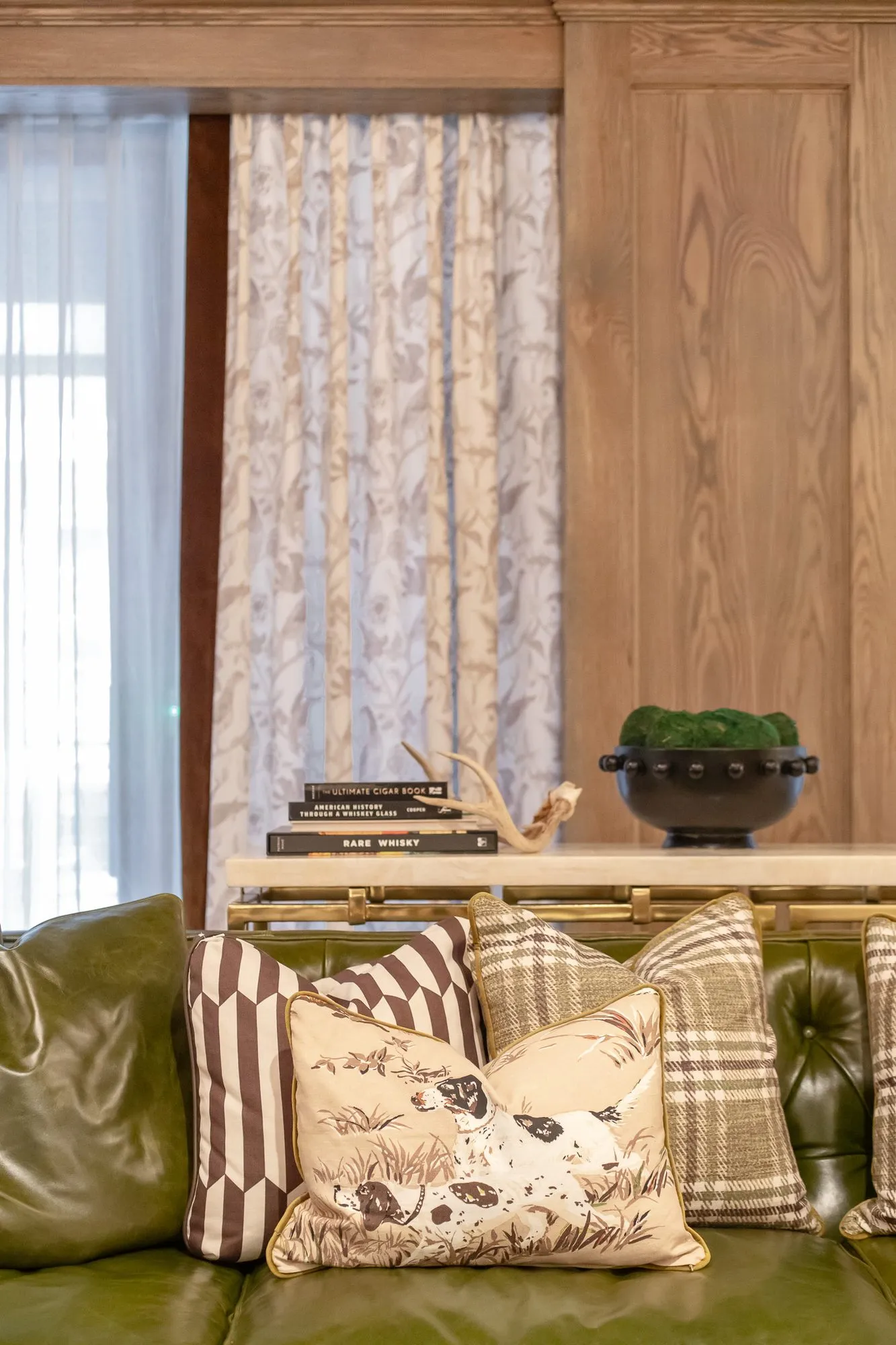 Private club interior detail: green tufted leather sofa with patterned pillows, a console with antlers and books by curtains