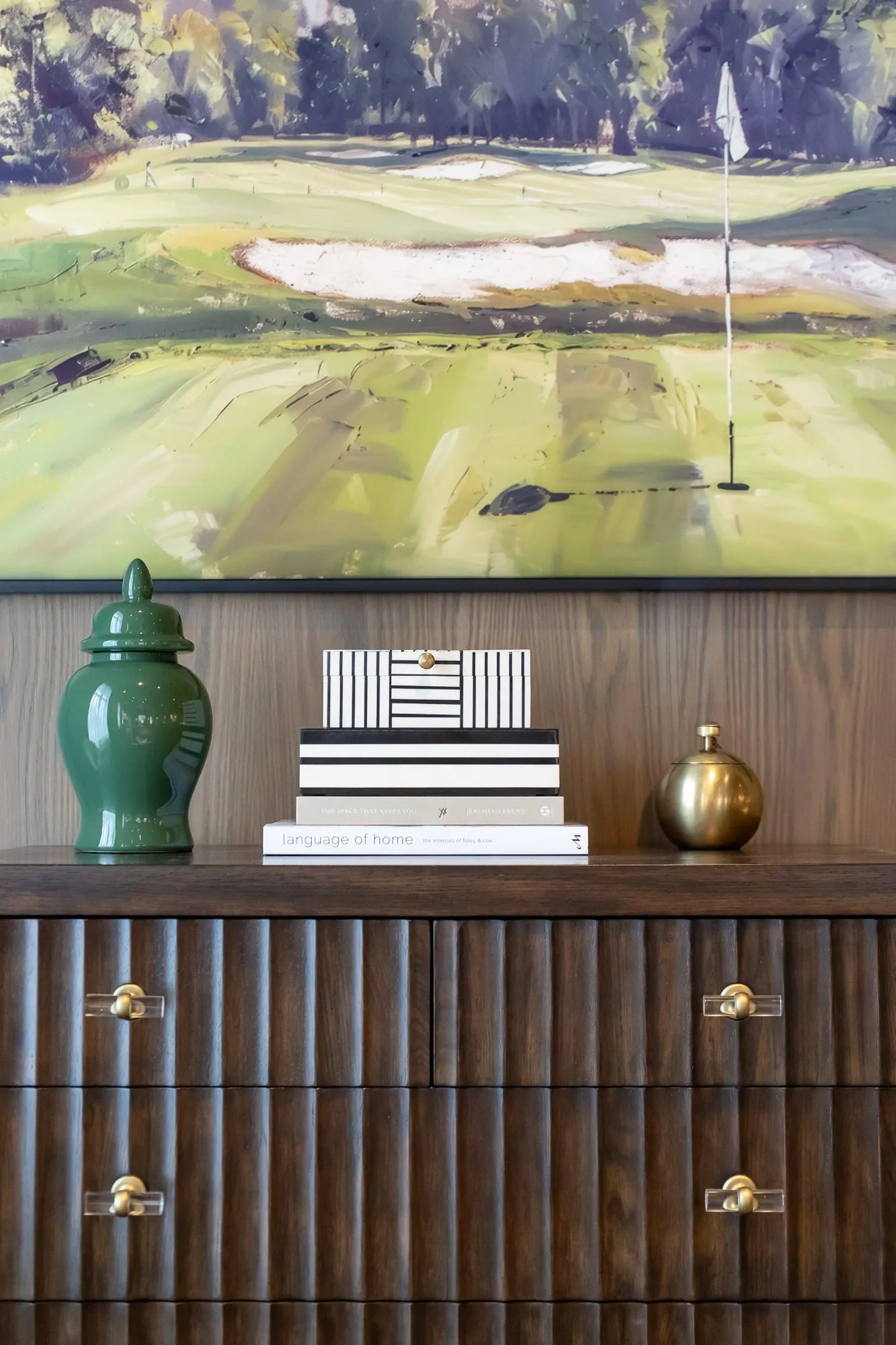 Private club interior detail: green ginger jar, striped boxes and a brass orb on a fluted walnut chest below a golf painting