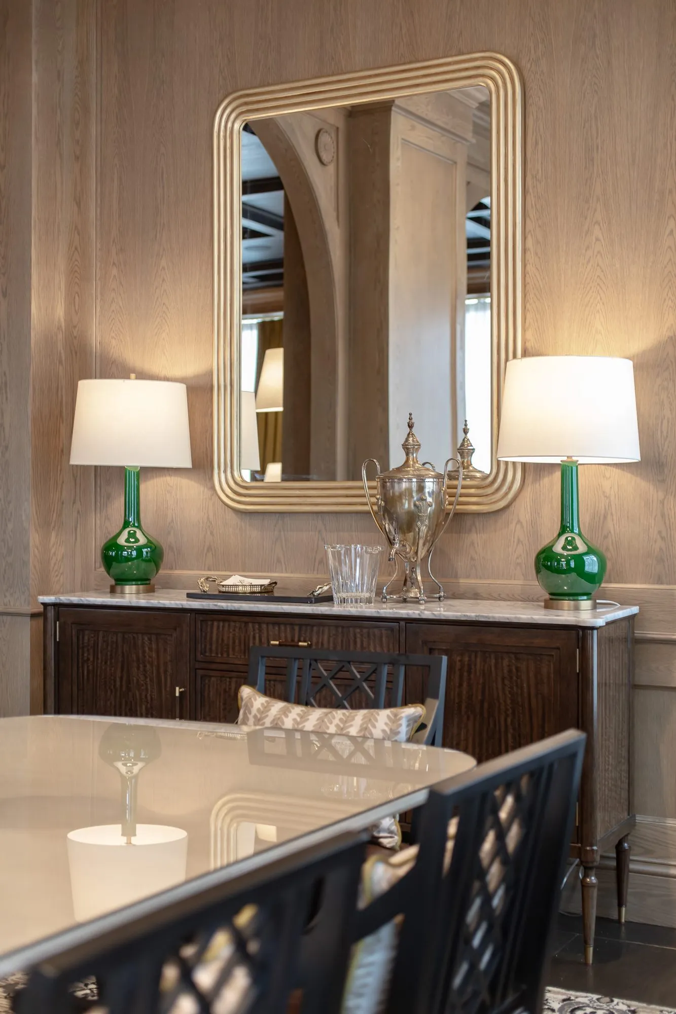 Private club dining room detail: gilt mirror over a marble sideboard with green glass lamps and a silver urn, chairs in front
