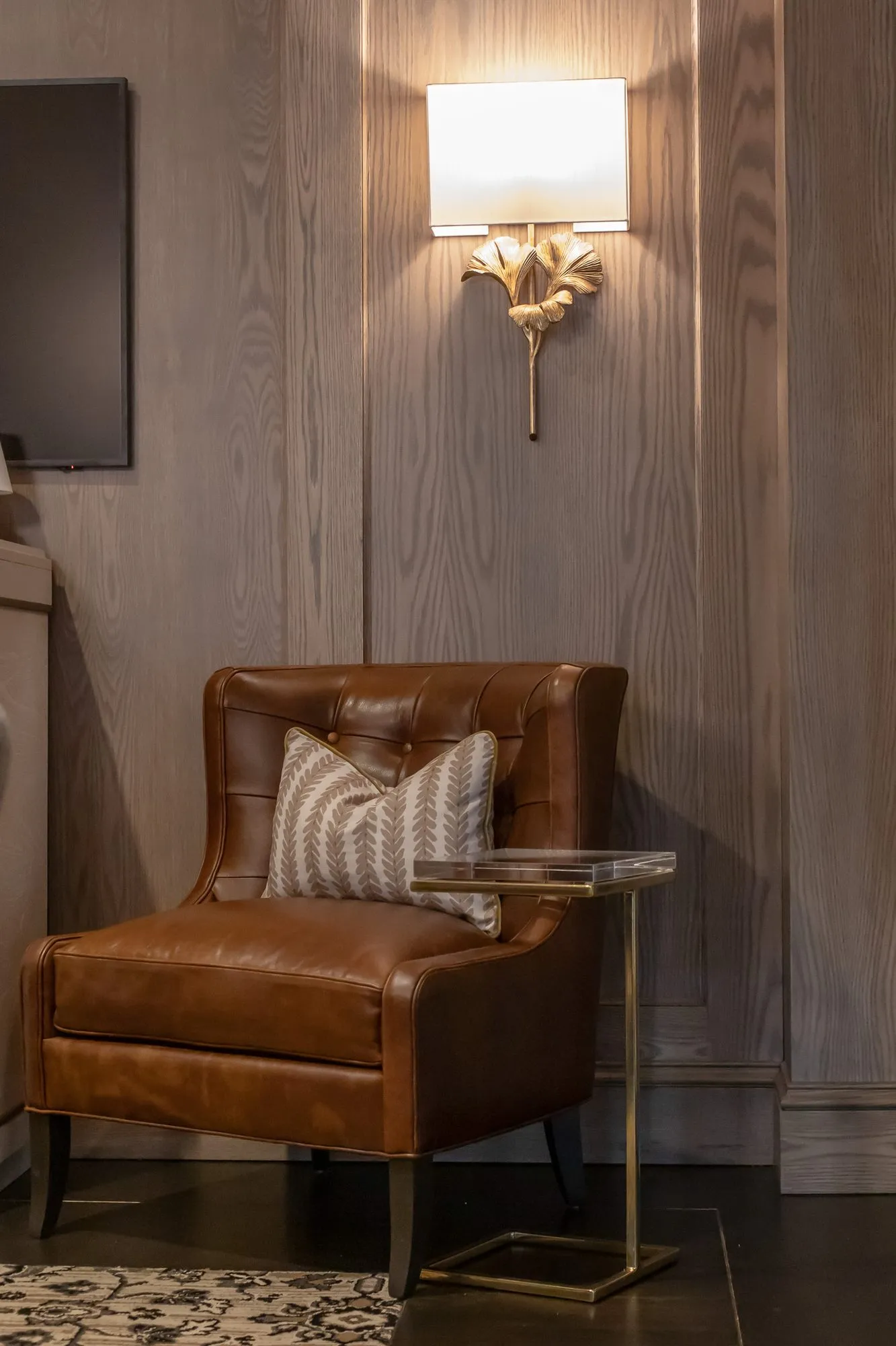 Private club interior detail: tan tufted leather chair with a striped pillow beside a brass side table under a ginkgo sconce