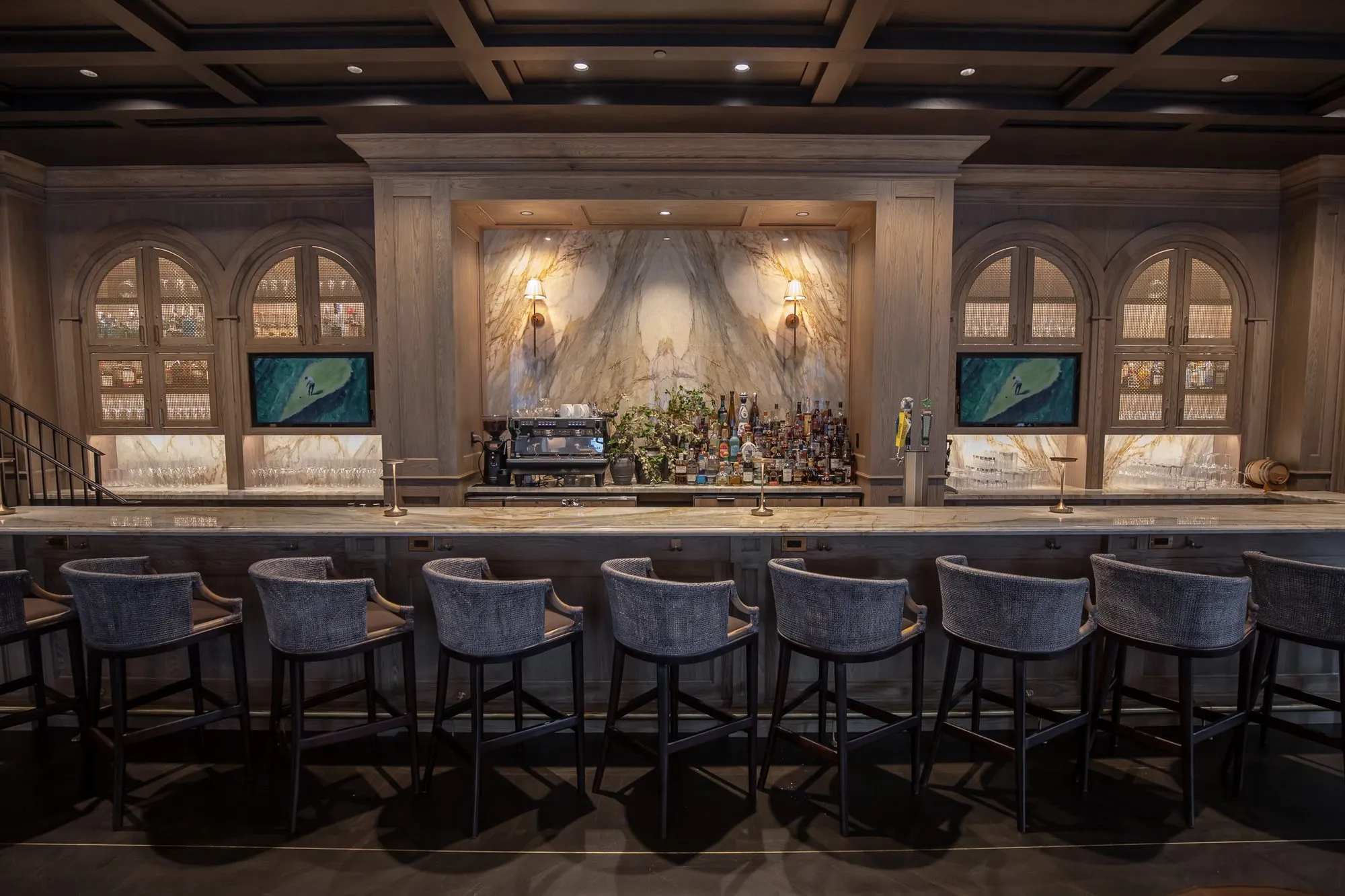 Private club bar with grey stools along the counter below the marble slab bar back and arched cabinets