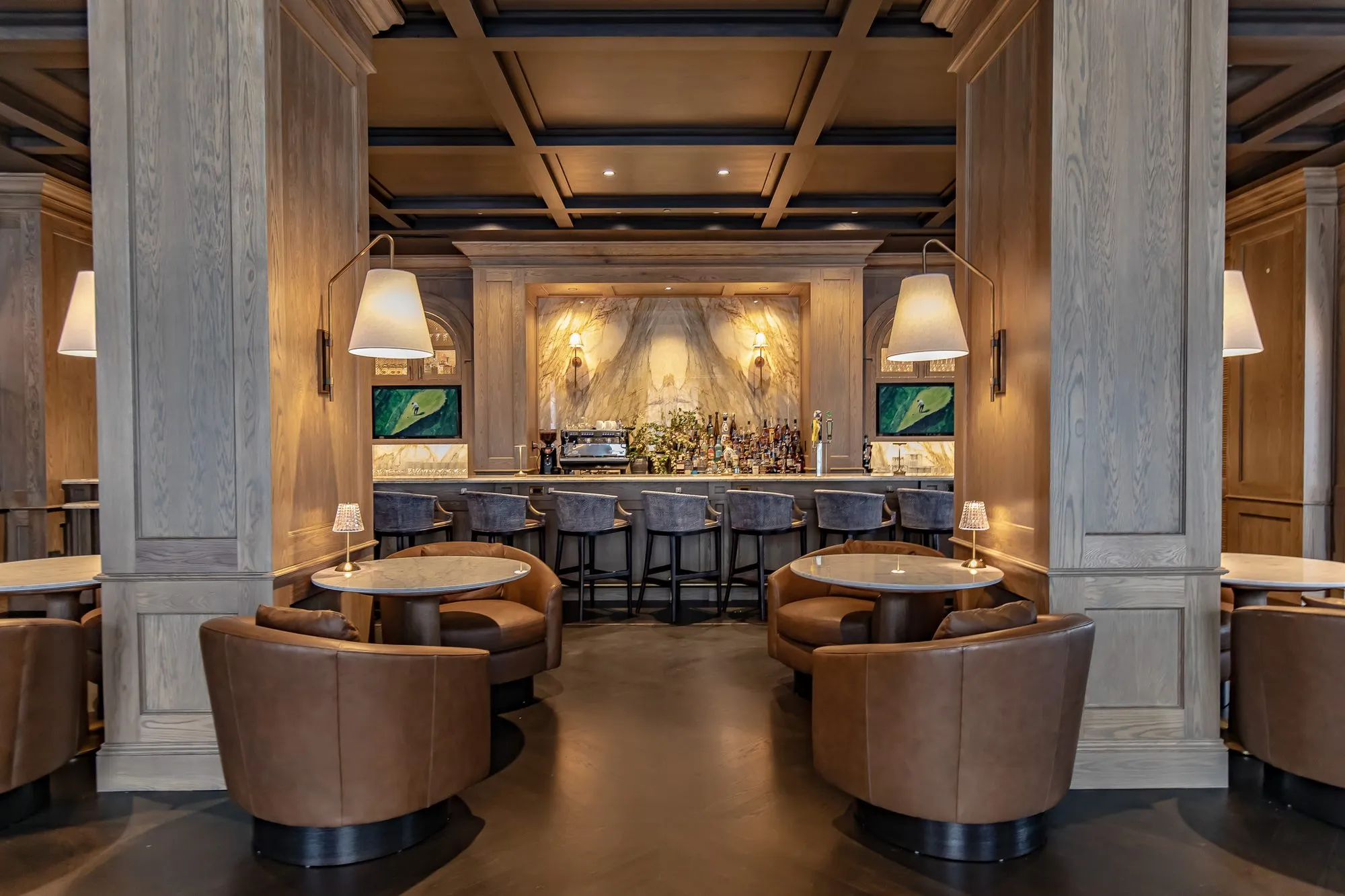 Private club bar seen between oak columns over paired leather tub chairs to the marble bar back