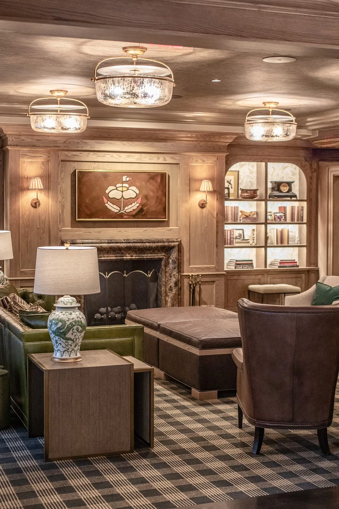 Private club library with a green leather sofa and ginger jar lamp, the crest painting over the fireplace and lit bookcase