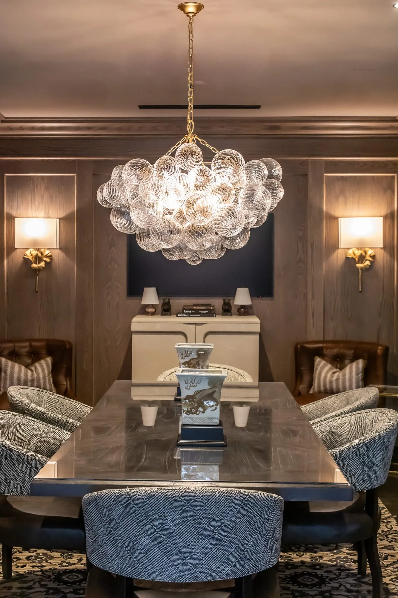 Private club dining room with grey patterned chairs at a long table under a bubble glass chandelier, gilt sconces on oak