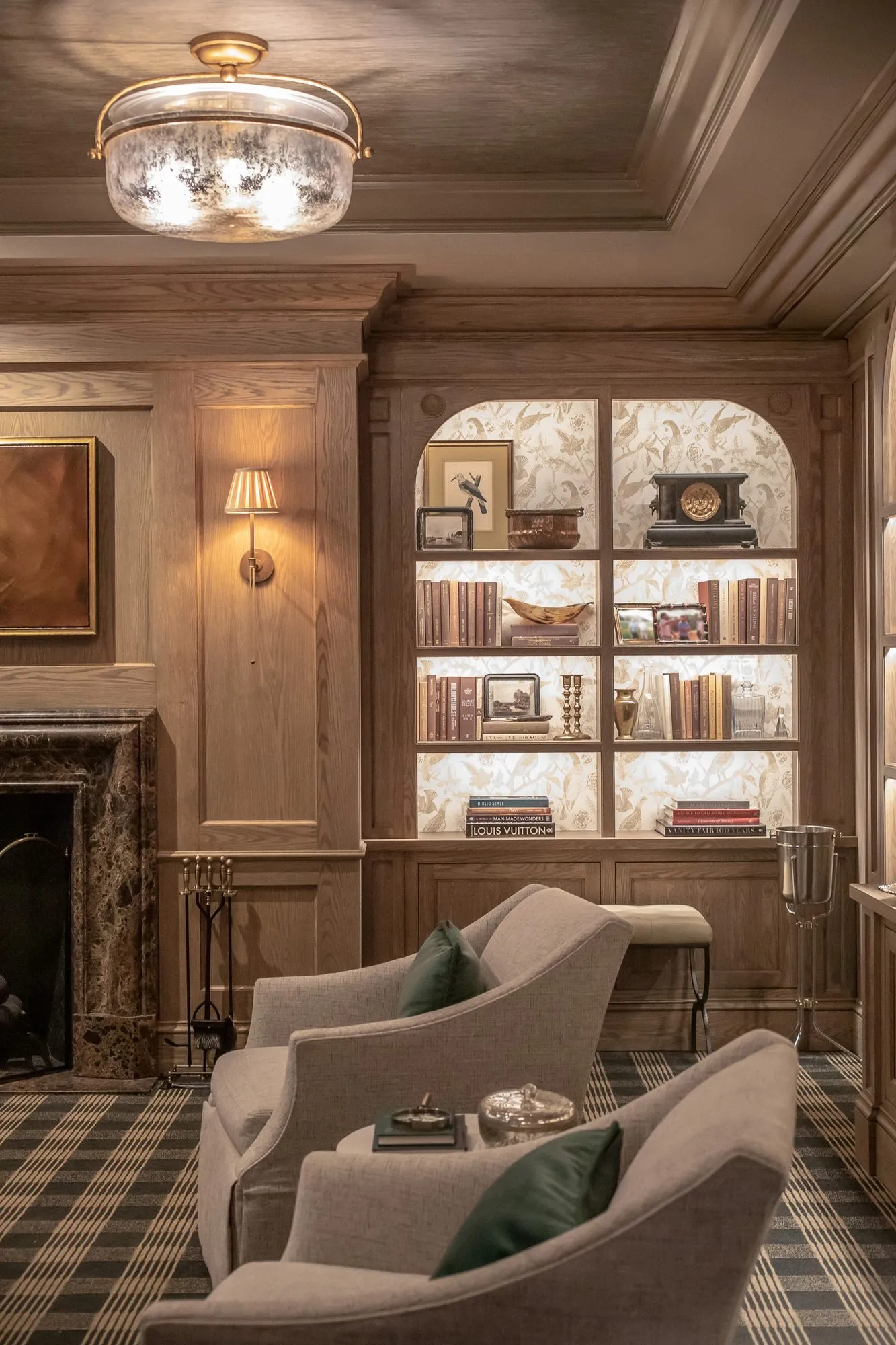 Private club interior detail: skirted armchairs with green pillows facing each other before a lit arched bookcase