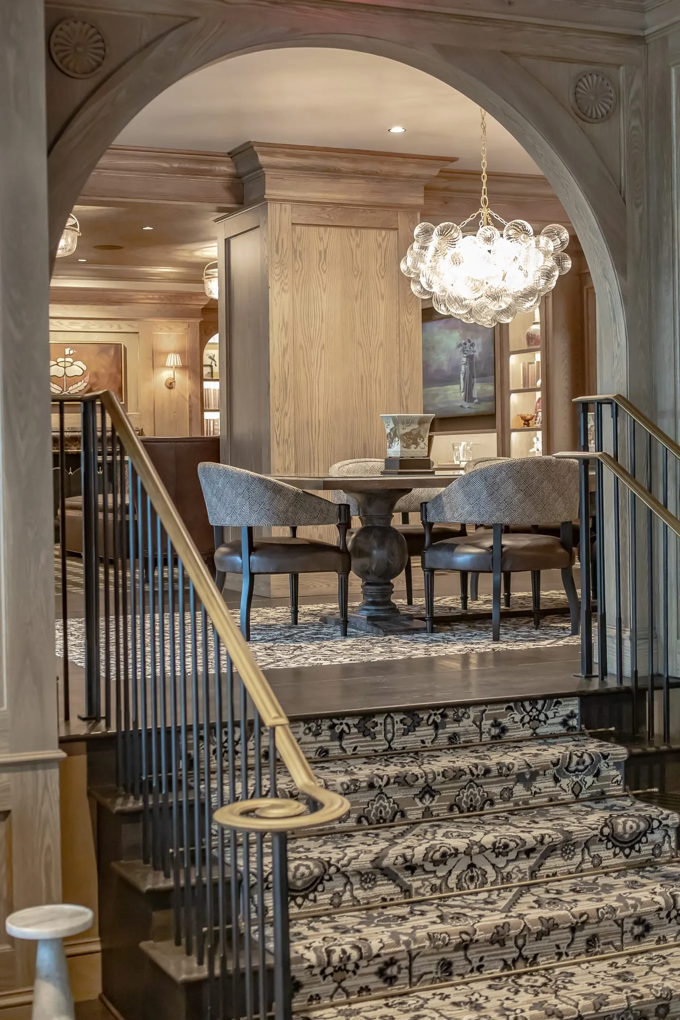 Private club staircase with a patterned runner and brass handrail rising through an arch to a round table, bubble chandelier