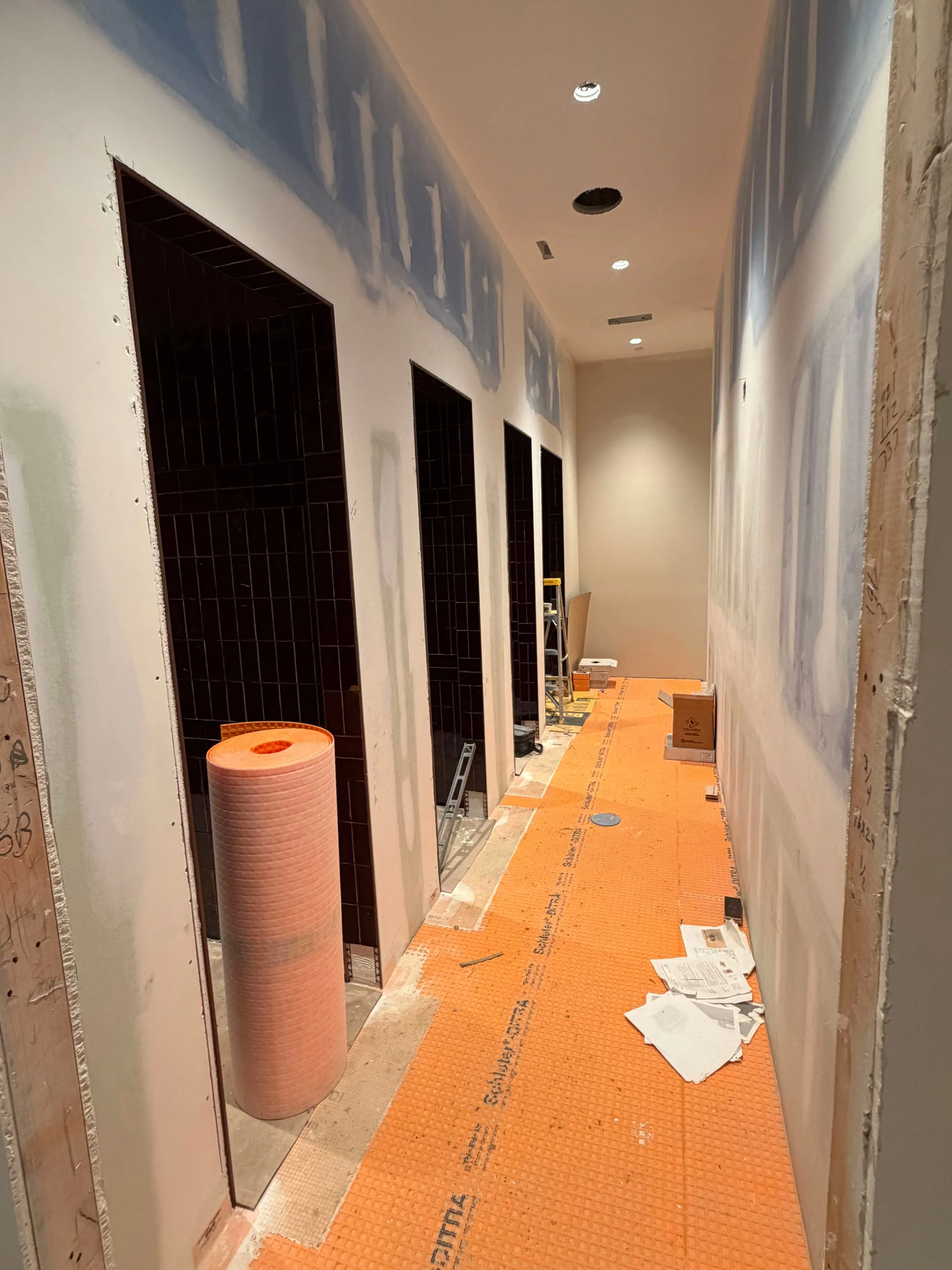 Private club clubhouse under construction: corridor of dark tiled shower stalls, orange waterproofing membrane on the floor