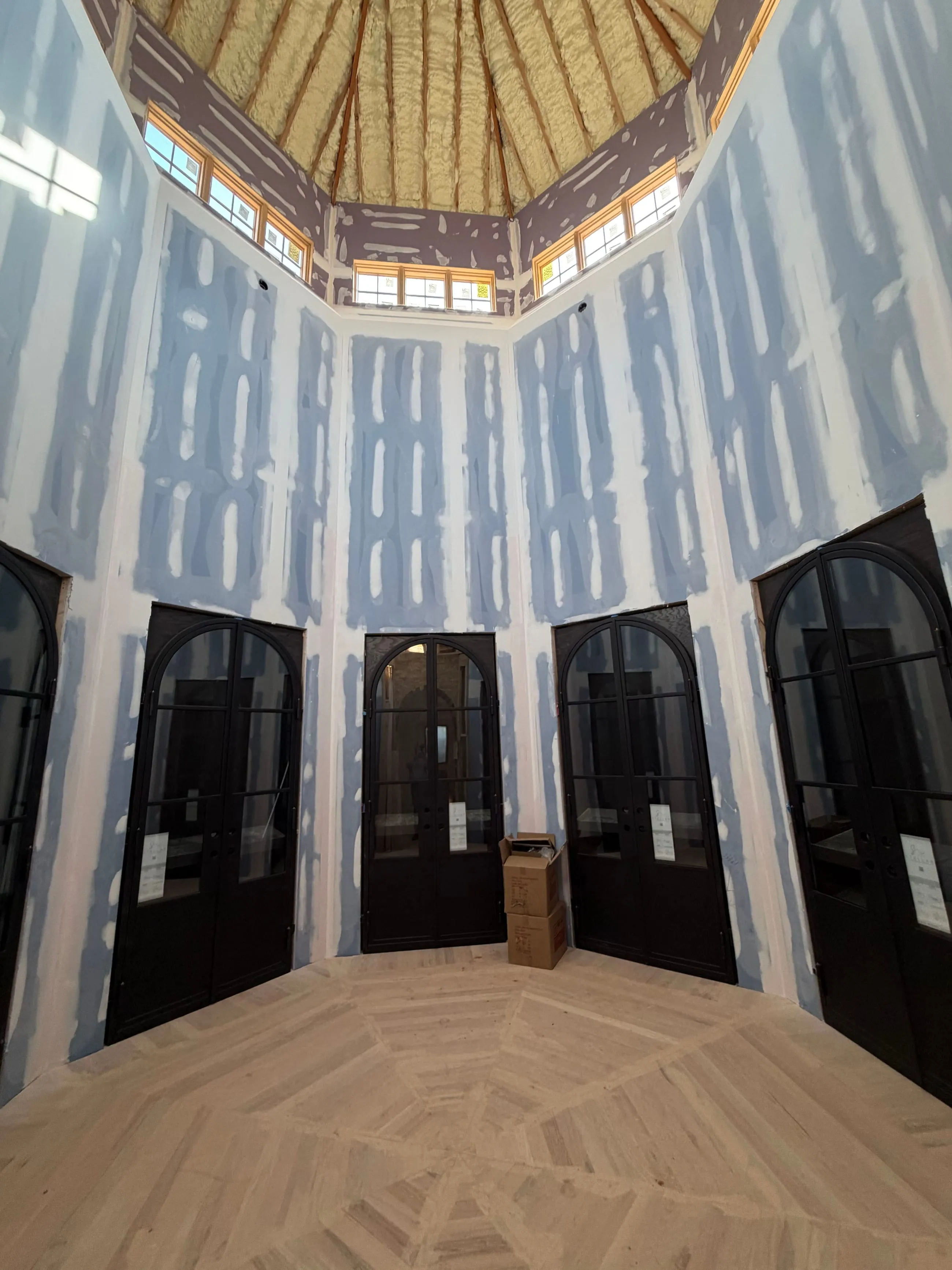 Private club clubhouse under construction: octagonal room of arched black steel glass doors in taped drywall, conical ceiling