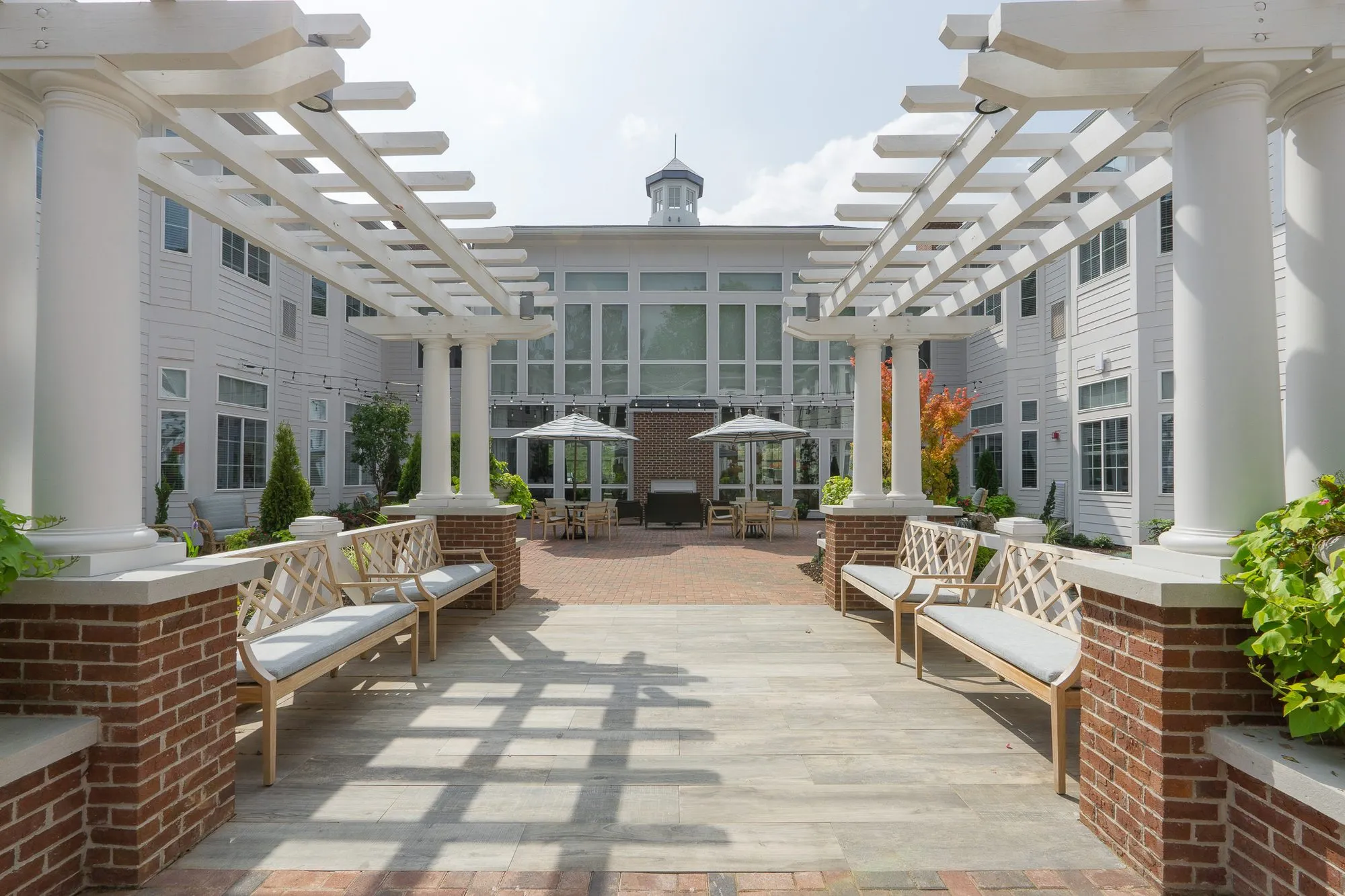 Senior living courtyard walk between brick planters and benches under twin white pergolas, a glass-walled wing with a cupola