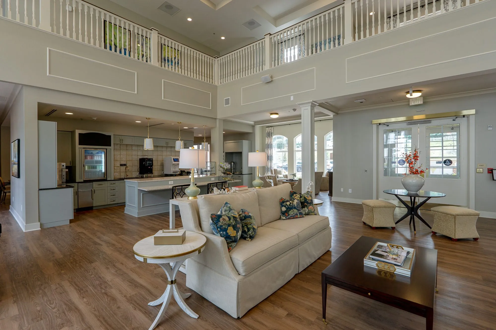 Senior living great room with a cream sofa and navy floral pillows below a second floor balcony, the open kitchen beyond