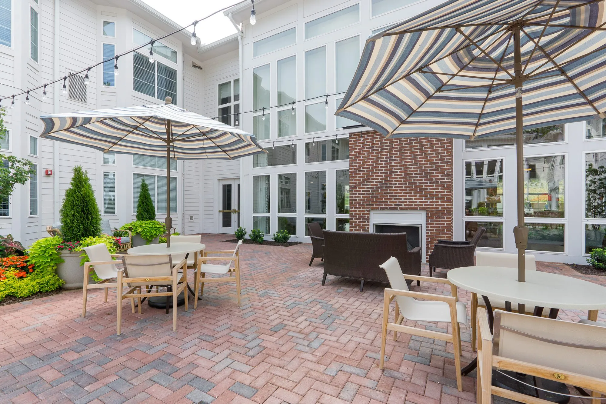 Senior living courtyard dining under striped umbrellas and string lights, a wicker sofa beside a brick outdoor fireplace