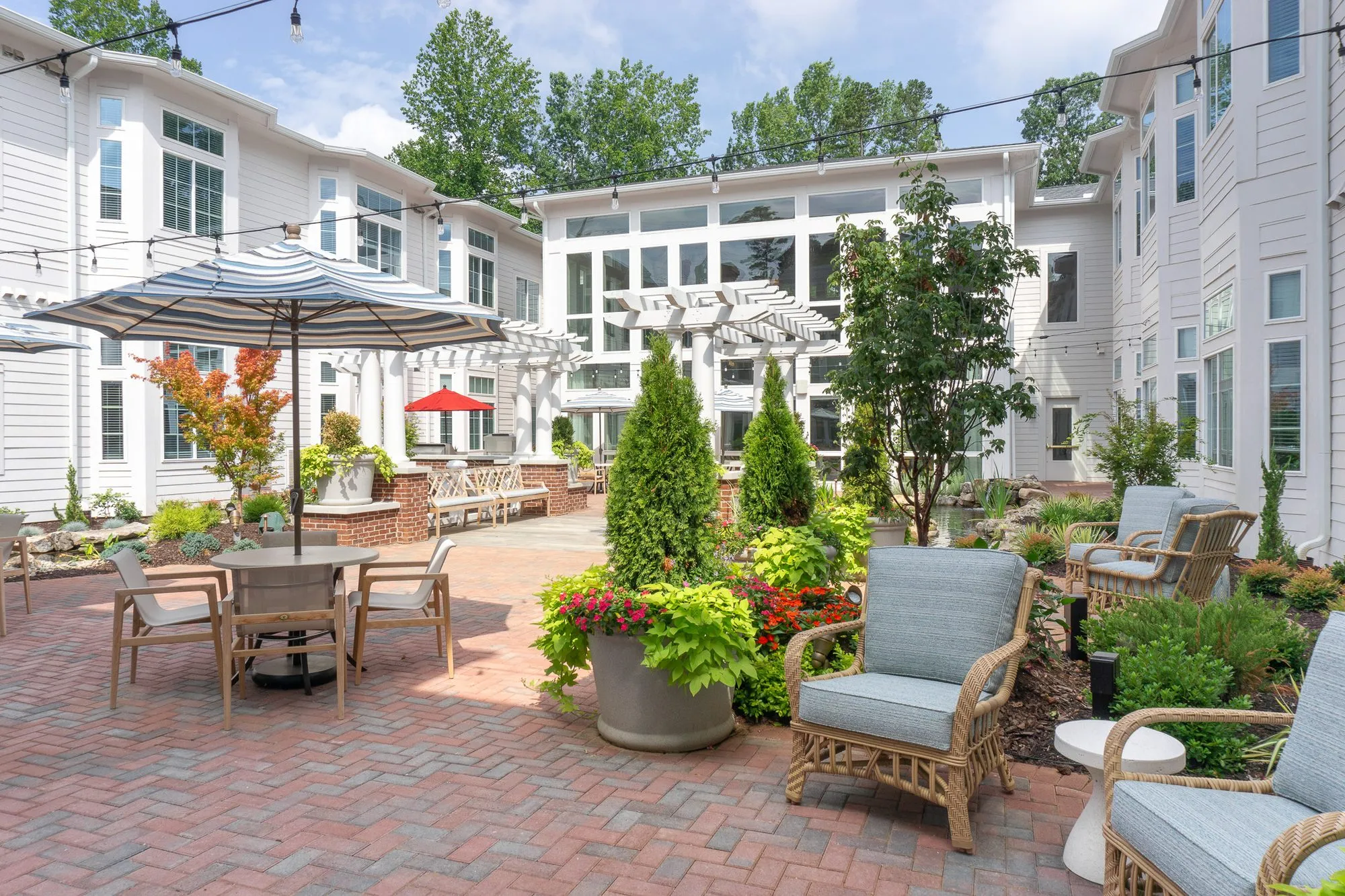 Senior living courtyard of herringbone brick with cypress and planted beds, a table under a striped umbrella and armchairs