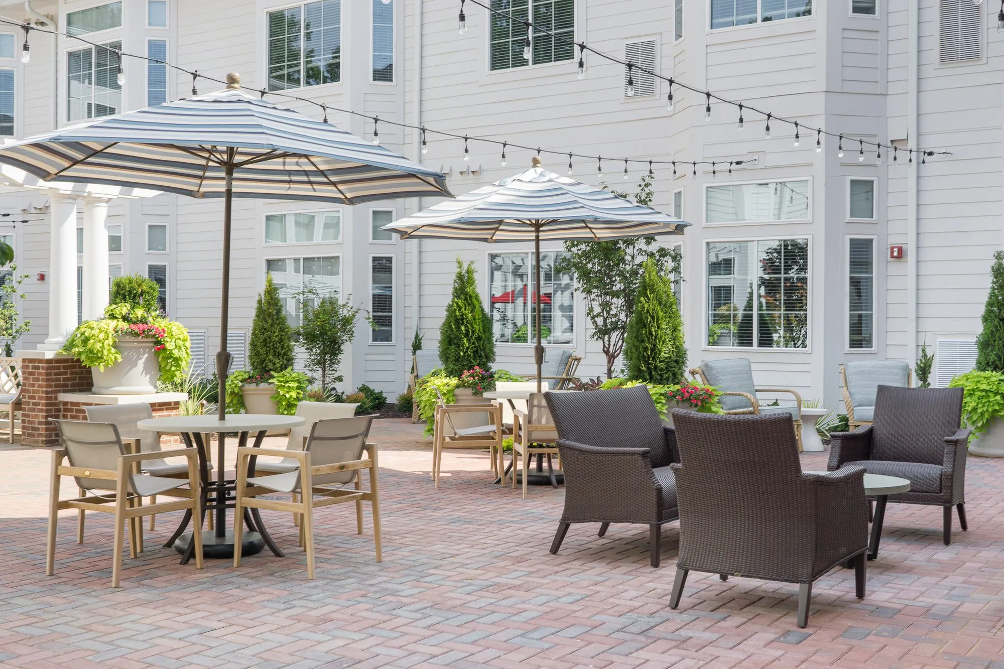 Senior living courtyard with string lights over a dining set and dark wicker lounge chairs between white clapboard walls