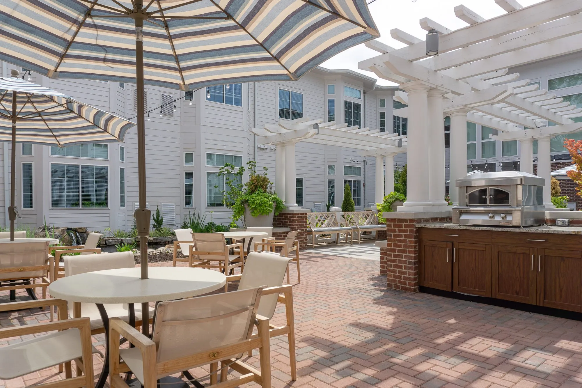 Senior living outdoor kitchen with wood cabinetry and a pizza oven under a white pergola, dining tables in front