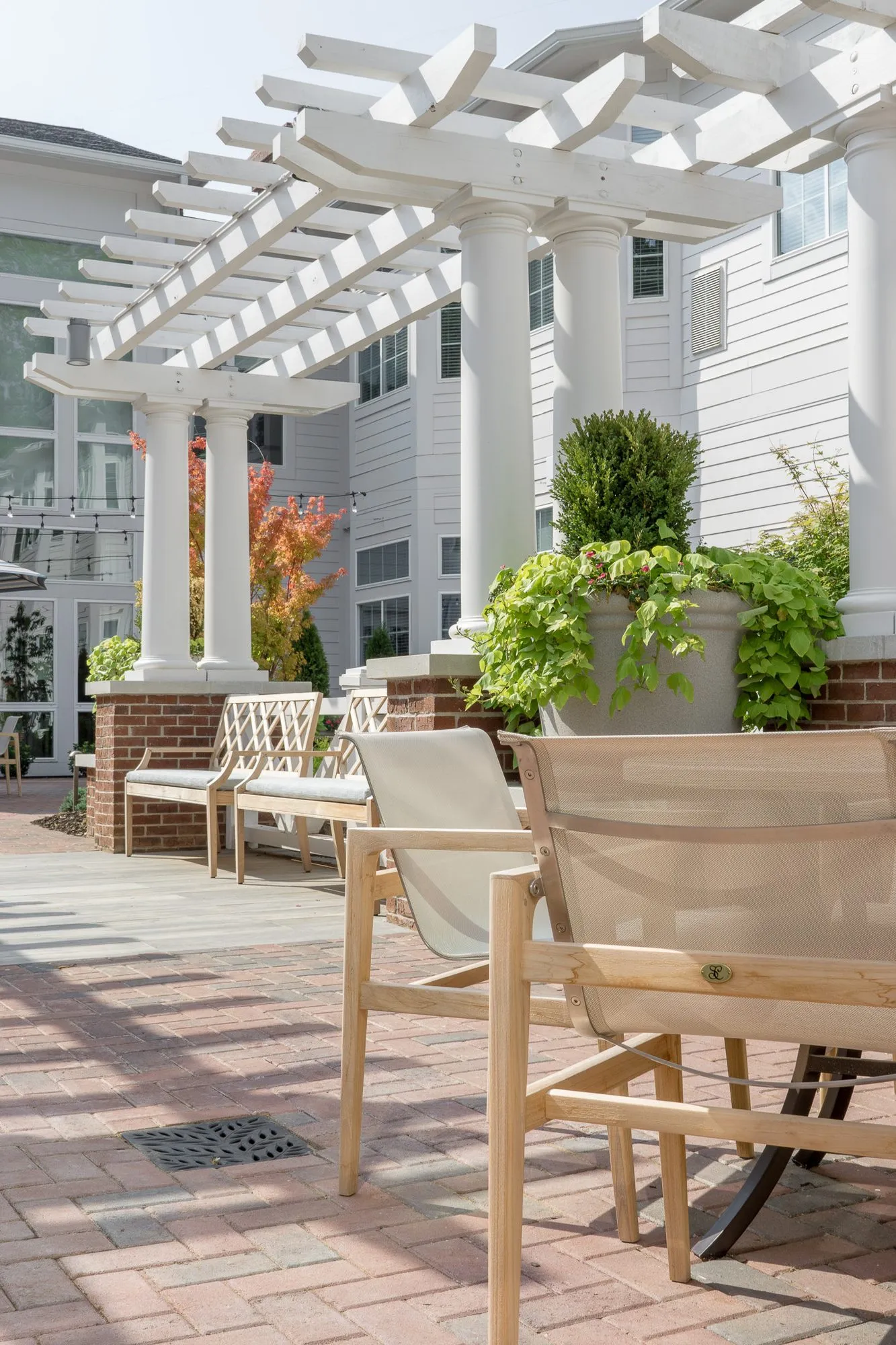 Senior living courtyard with a white pergola over brick planters and benches, a teak chair in the foreground