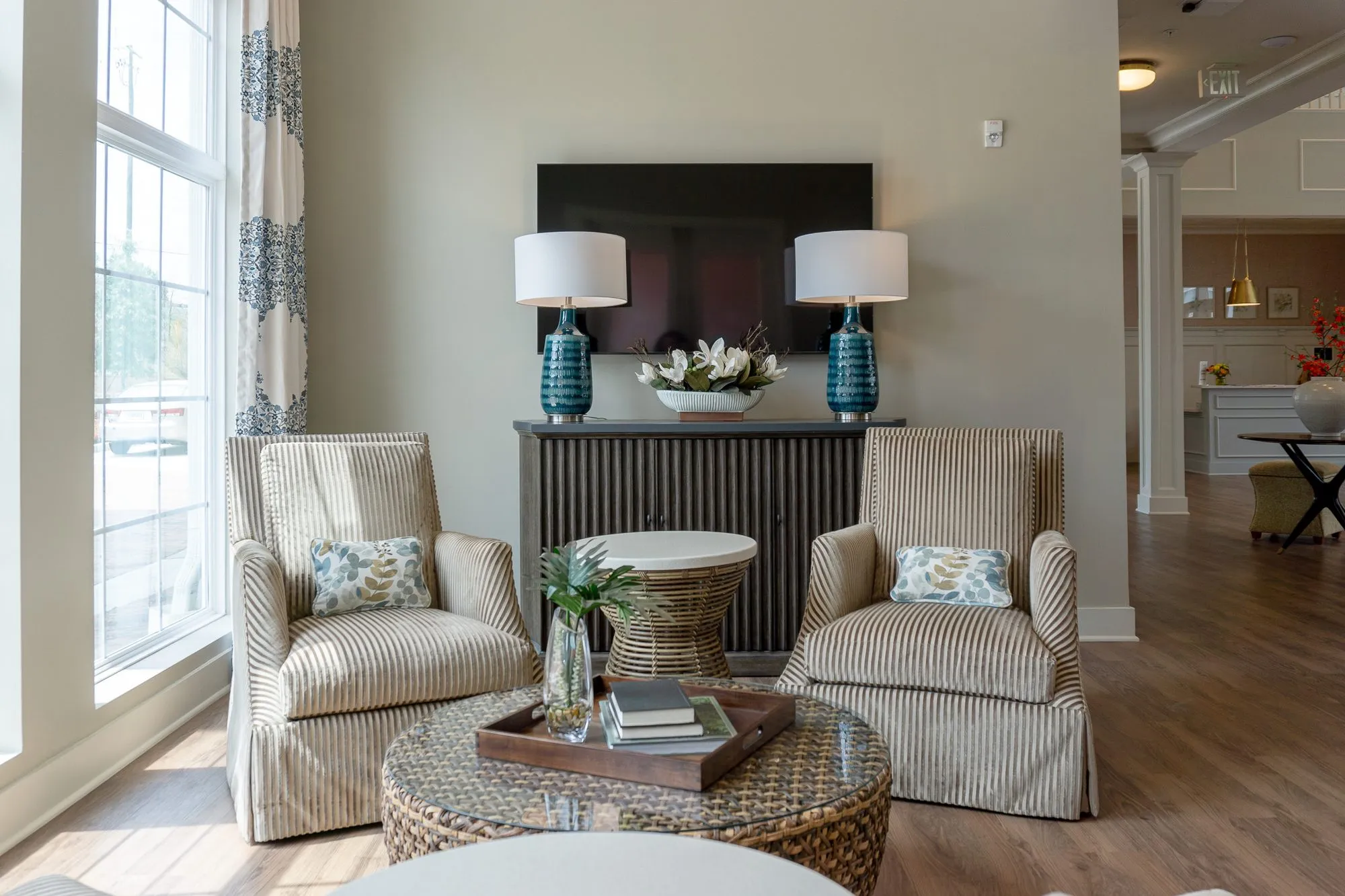 Senior living sitting room with striped skirted armchairs at a woven coffee table by tall windows, teal lamps behind
