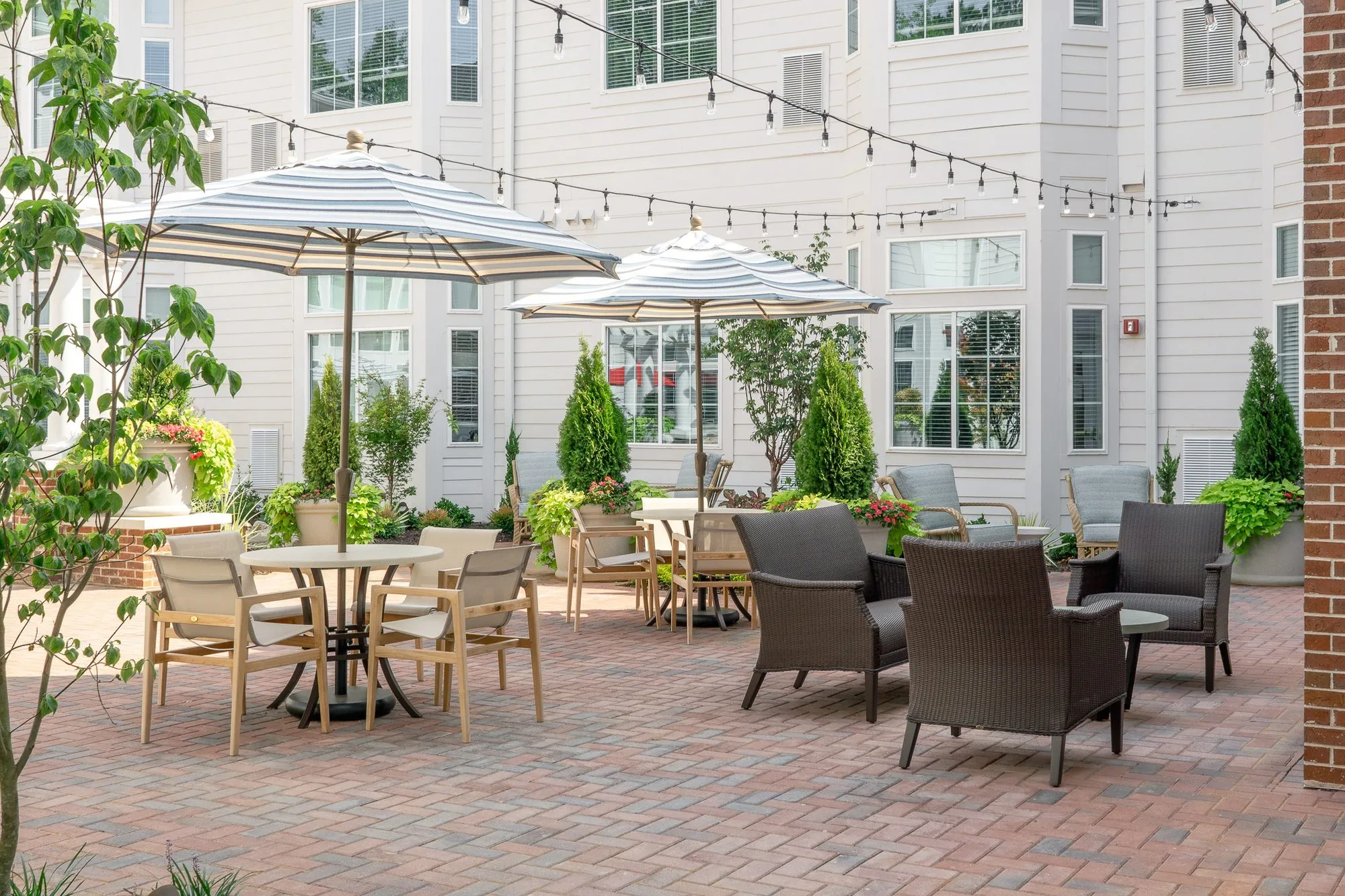 Senior living courtyard corner with two dining tables under striped umbrellas and a group of dark wicker armchairs