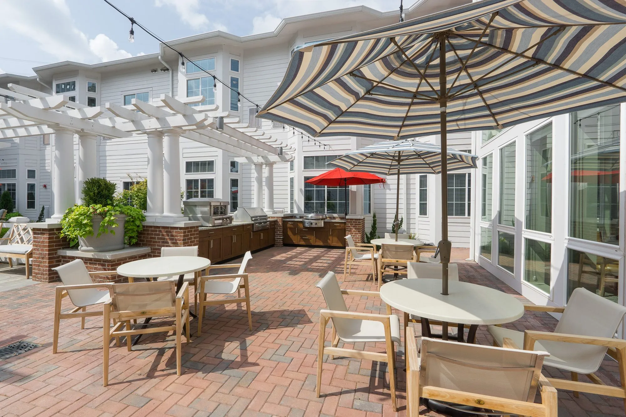 Senior living courtyard dining tables under a striped umbrella, a white pergola over a brick planter and the outdoor kitchen