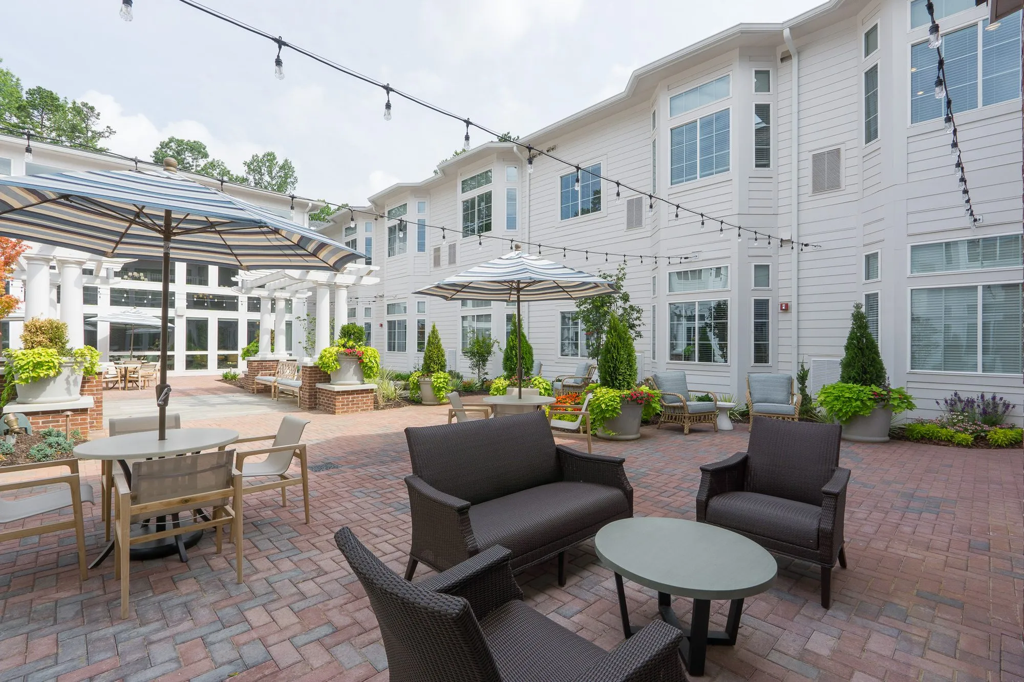 Senior living courtyard with a dark wicker sofa group in front and striped umbrellas along string-lit walls