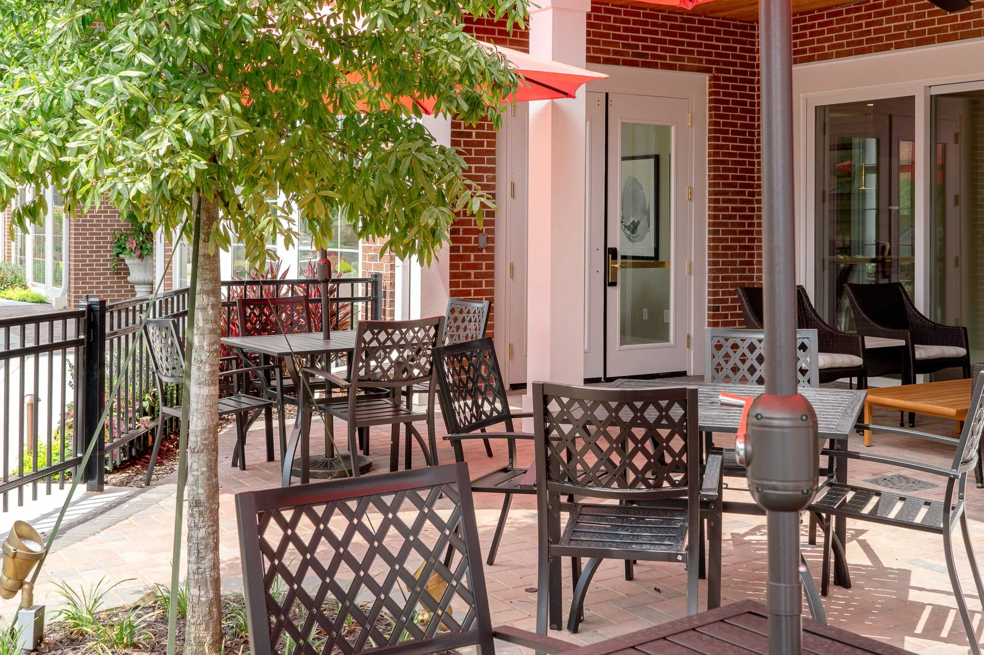 Senior living terrace with dark metal lattice chairs at patio tables on fenced brick beside a red door