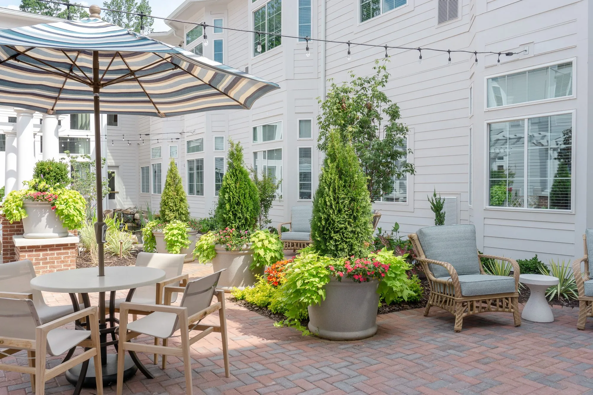 Senior living courtyard dining table under a striped umbrella beside cypress and annuals, teak armchairs by the clapboard